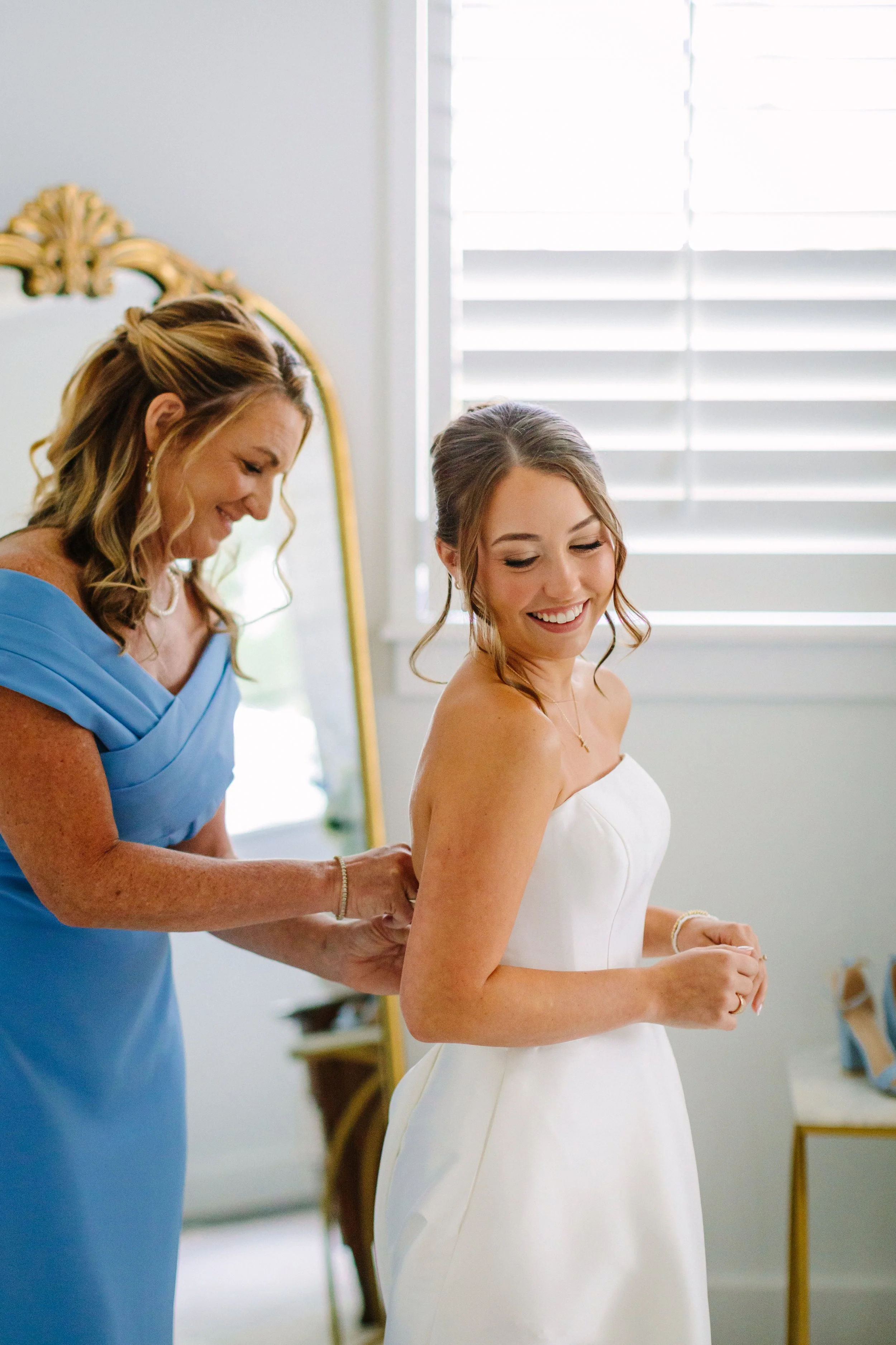 A woman in a white dress, likely a bride, smiling as another woman, possibly her mother or a maid of honor, helps her adjust her dress in a well-lit room with white shutters.