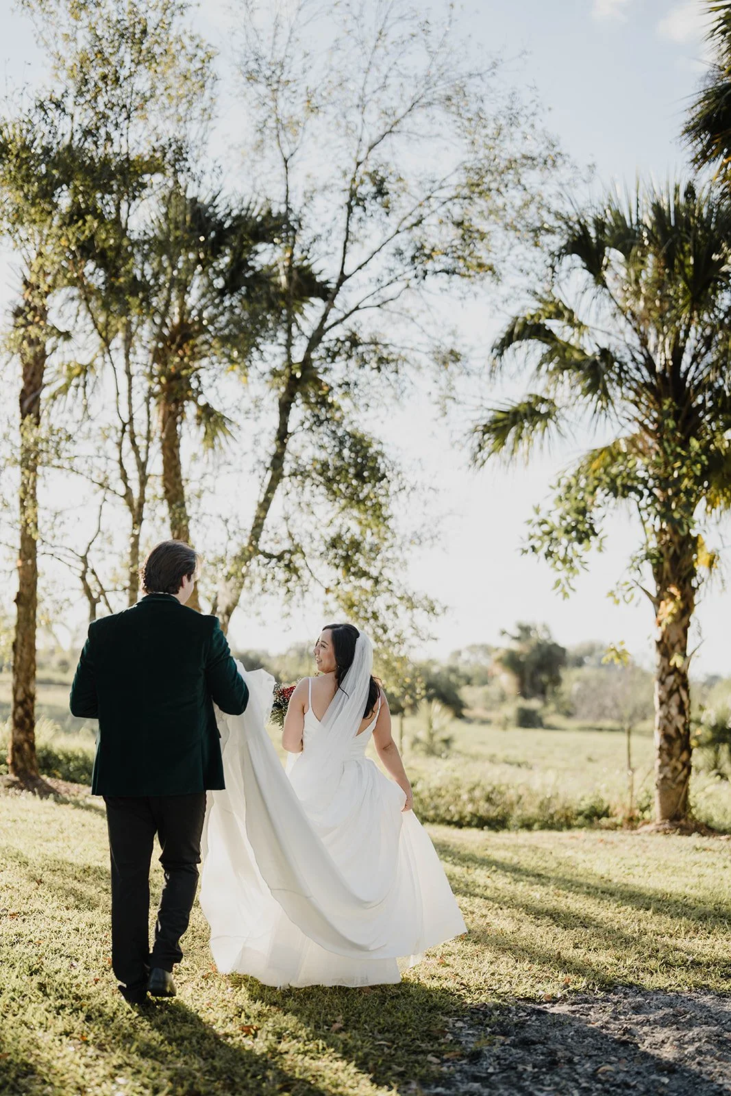 A bride and groom walking outdoors on a sunny day, with trees and a grassy landscape in the background. The bride wears a white wedding dress and veil, and the groom is dressed in a black suit.