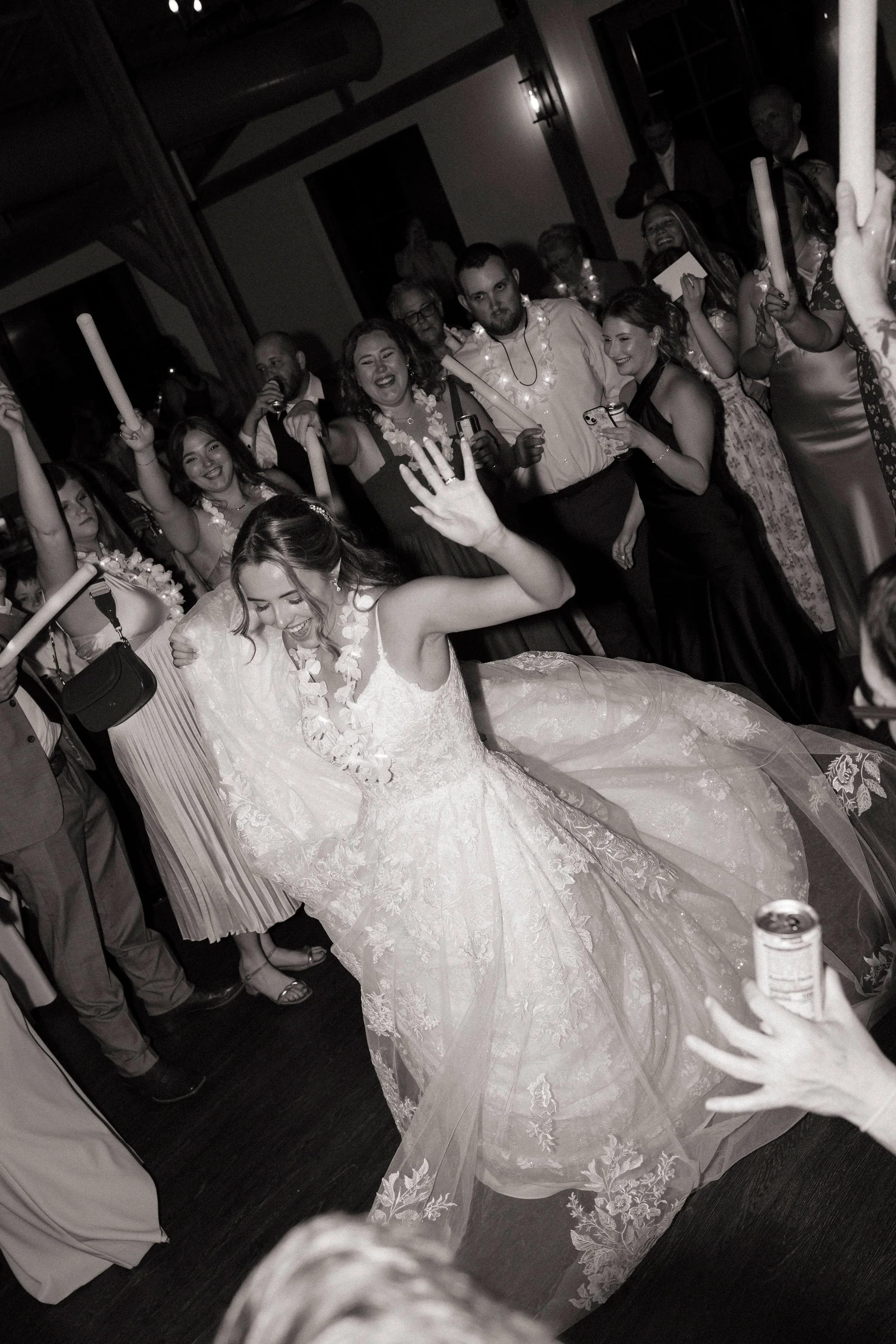 Bride dancing at her wedding reception with guests surrounding her, some holding sticks or drinks, in a decorated indoor venue.
