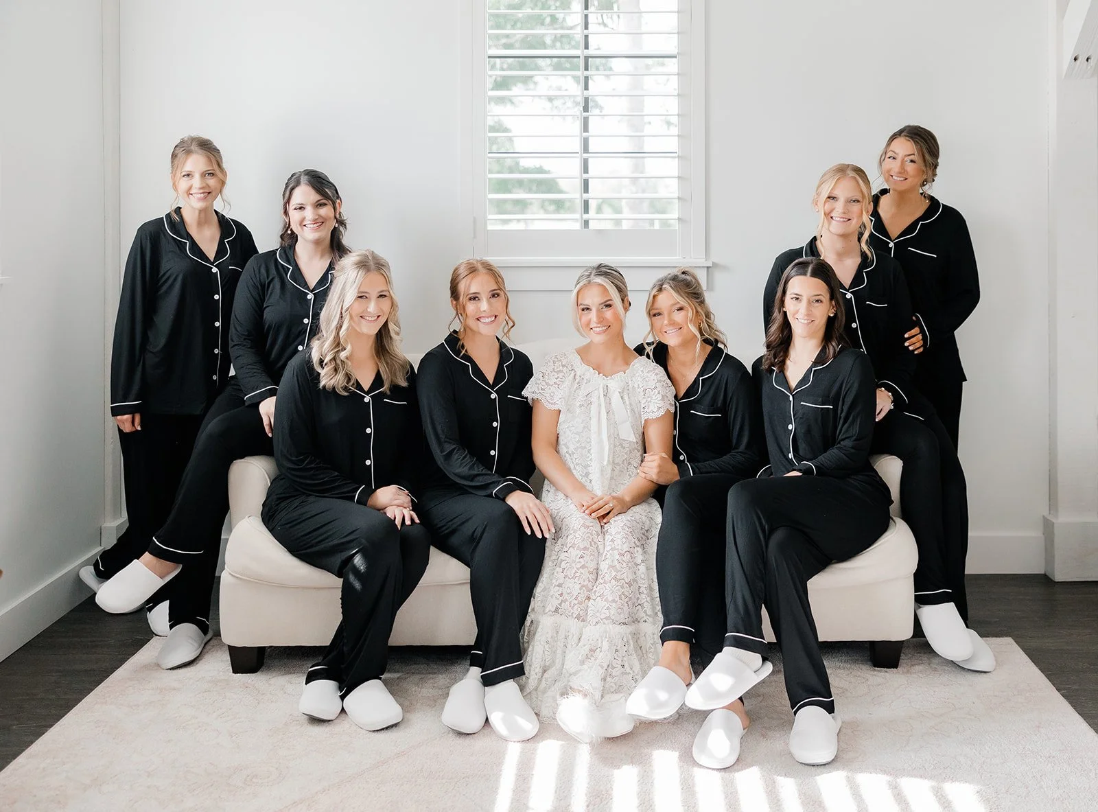 A bride in a white lace dress and nine bridesmaids in matching black pajamas with white piping, all wearing white slippers, sitting on and around a white sofa in a bright room with a window.