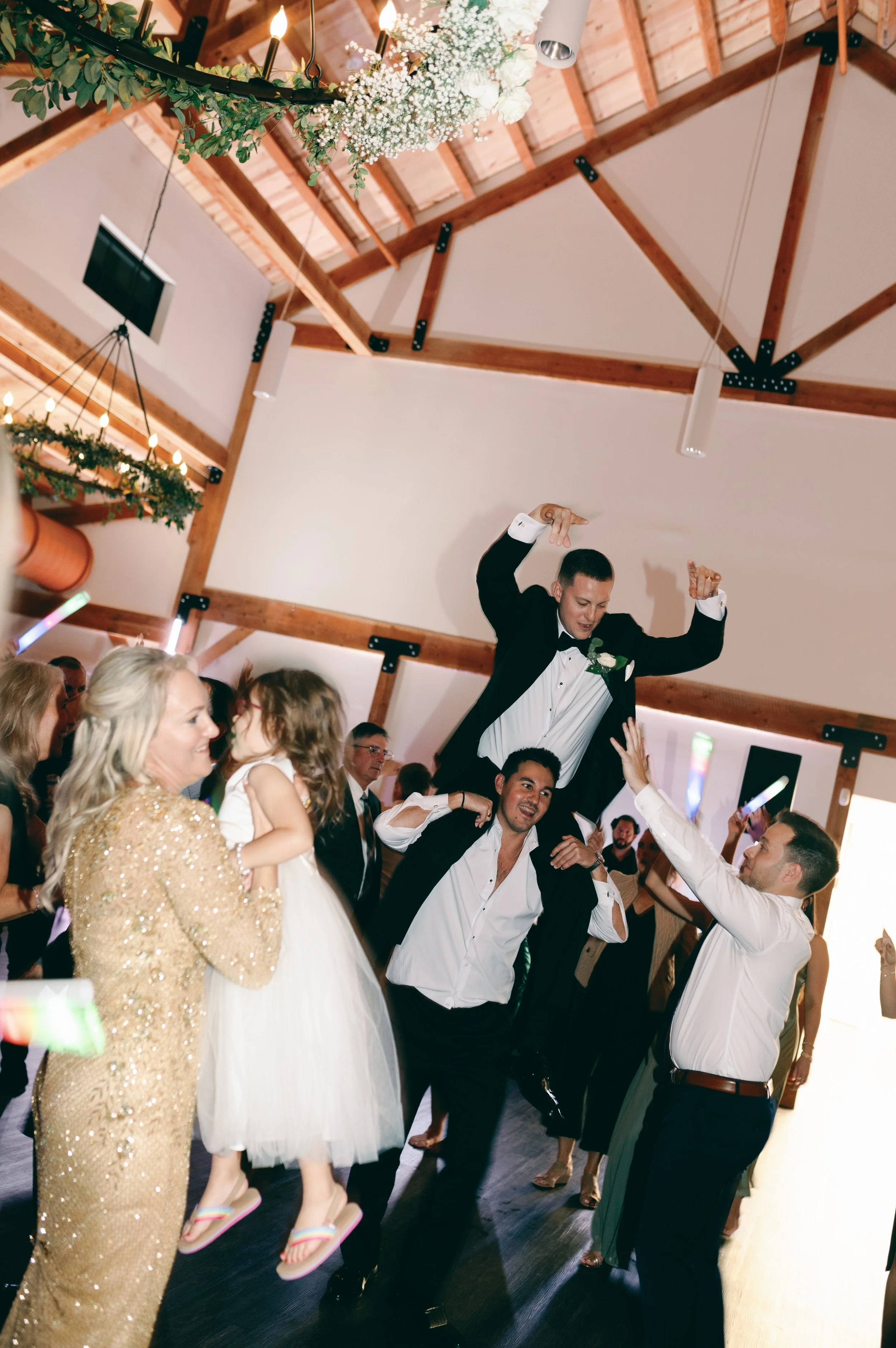 A group of people celebrating at a wedding reception, with a groom lifted above others, wearing a tuxedo and smiling, while others dance and watch.