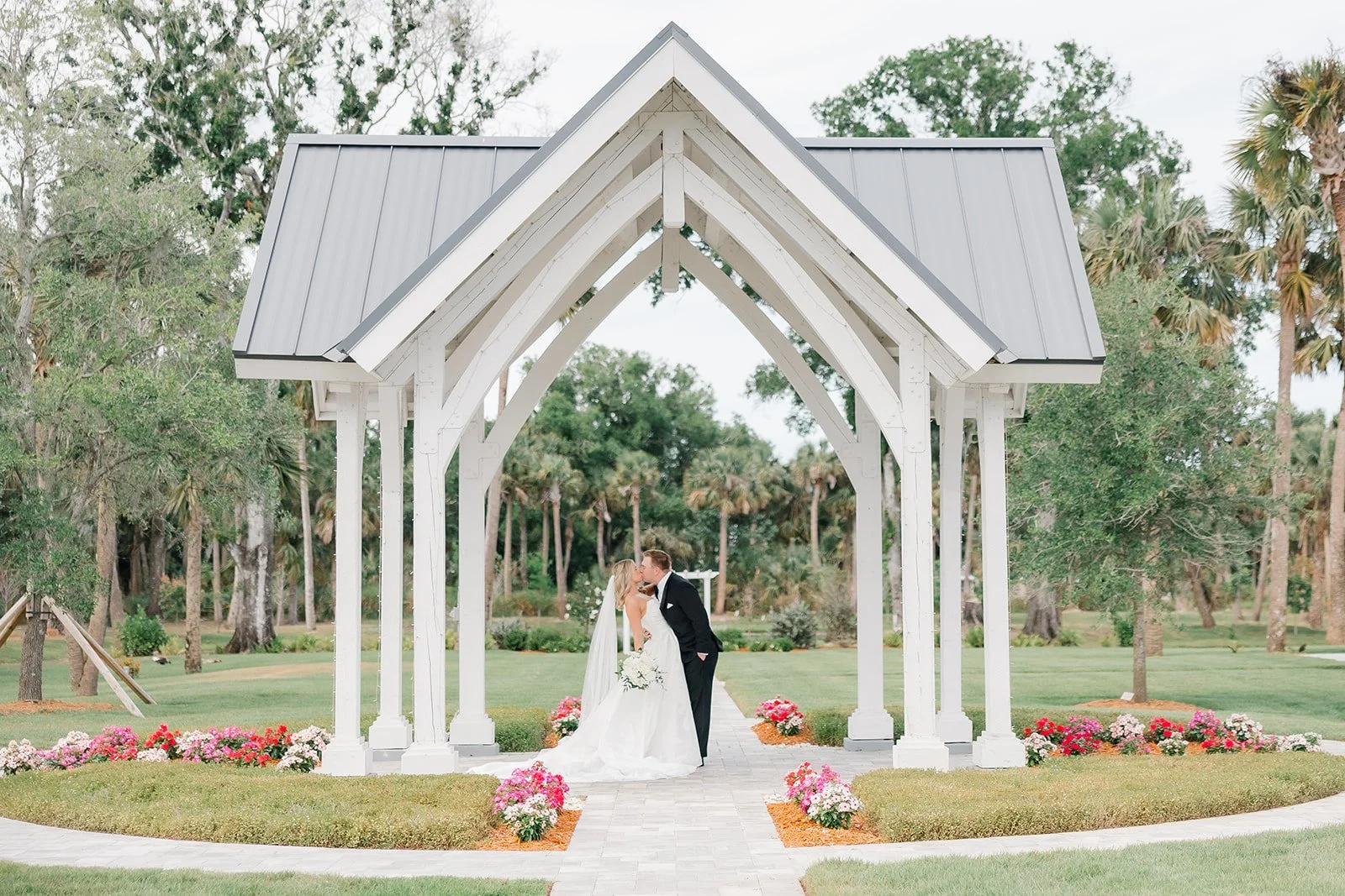 A newlywed couple kissing under a white wooden pavilion during a wedding ceremony outdoor with green trees and flowers.
