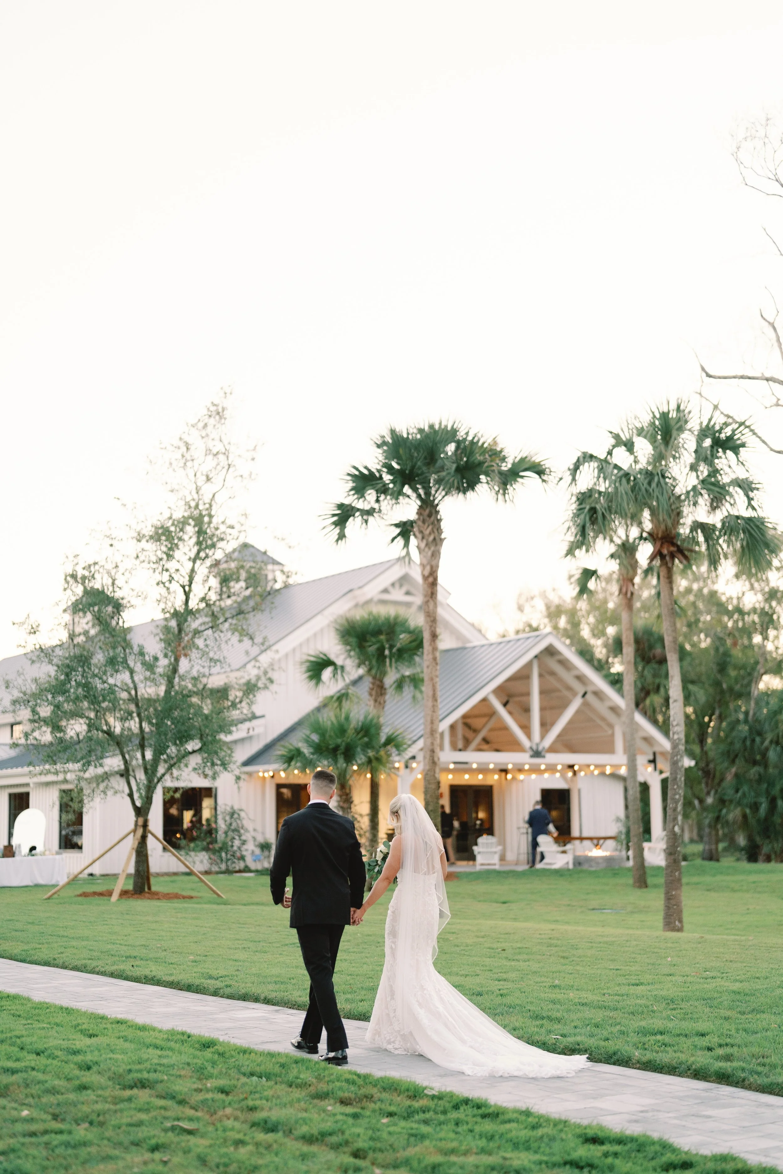 A bride and groom walking hand in hand on a stone path in a grassy outdoor area at a wedding reception, with a white barn-style building and palm trees in the background during sunset.