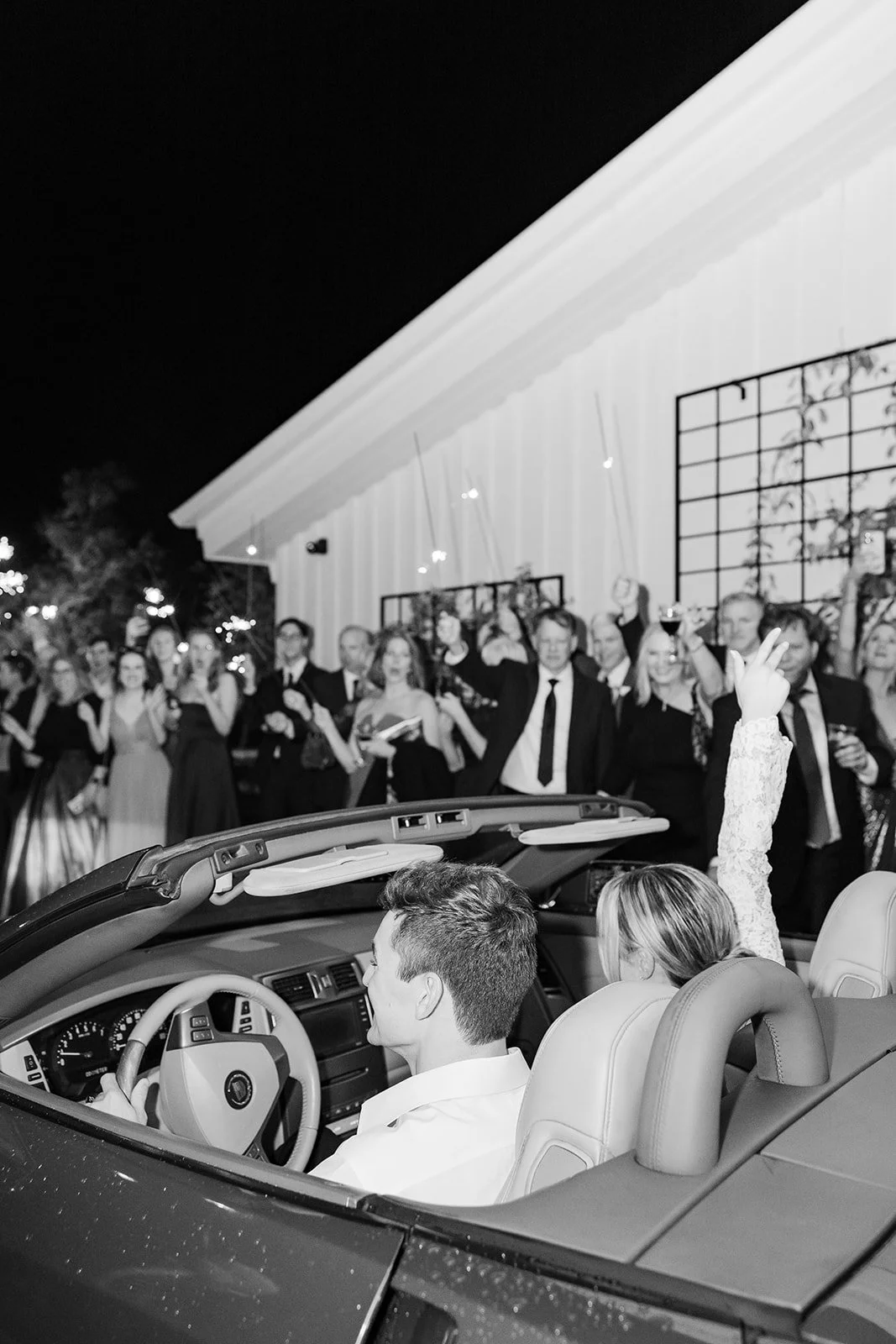 Black and white photo of a wedding celebration at night with guests raising glasses, standing outside a house with lights, as a newlywed couple sits in a convertible car in front of them.