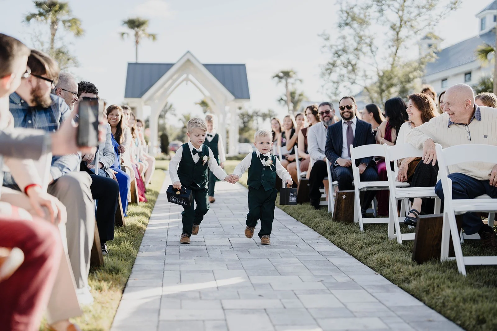 Two young boys dressed in formal suits walking down an outdoor aisle at a wedding, holding hands and carrying small suitcases labeled 'Security', surrounded by seated guests on either side.