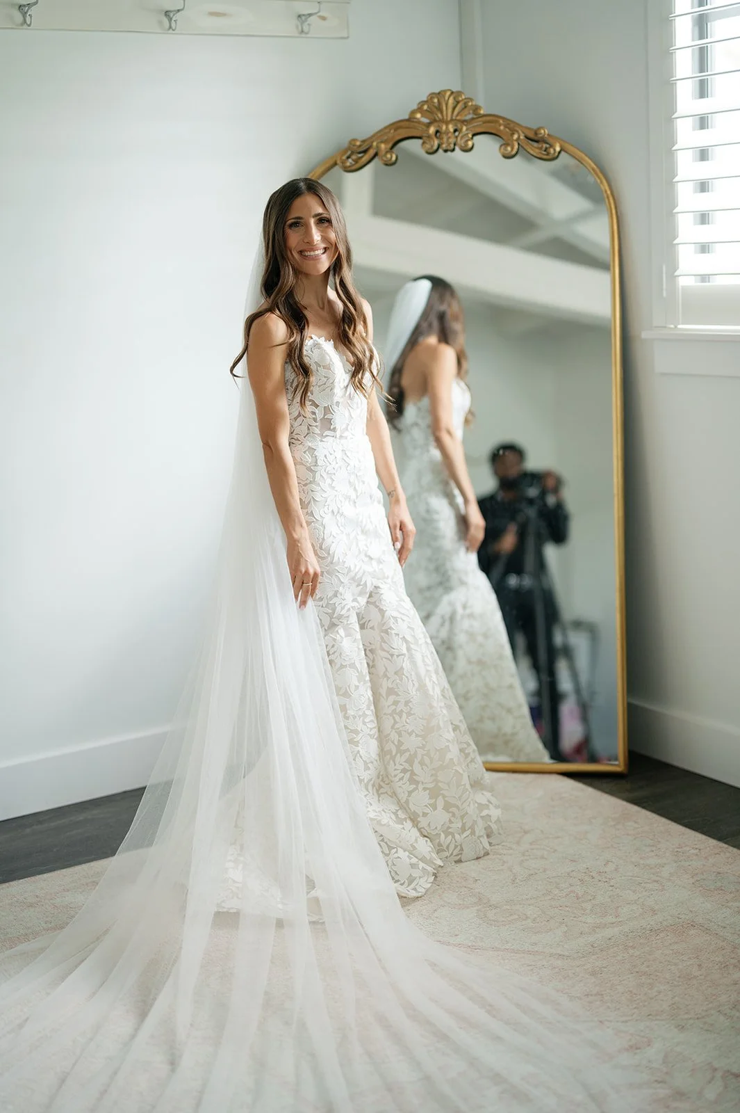 A smiling woman in a wedding dress with a veil standing in front of a mirror in a well-lit room.
