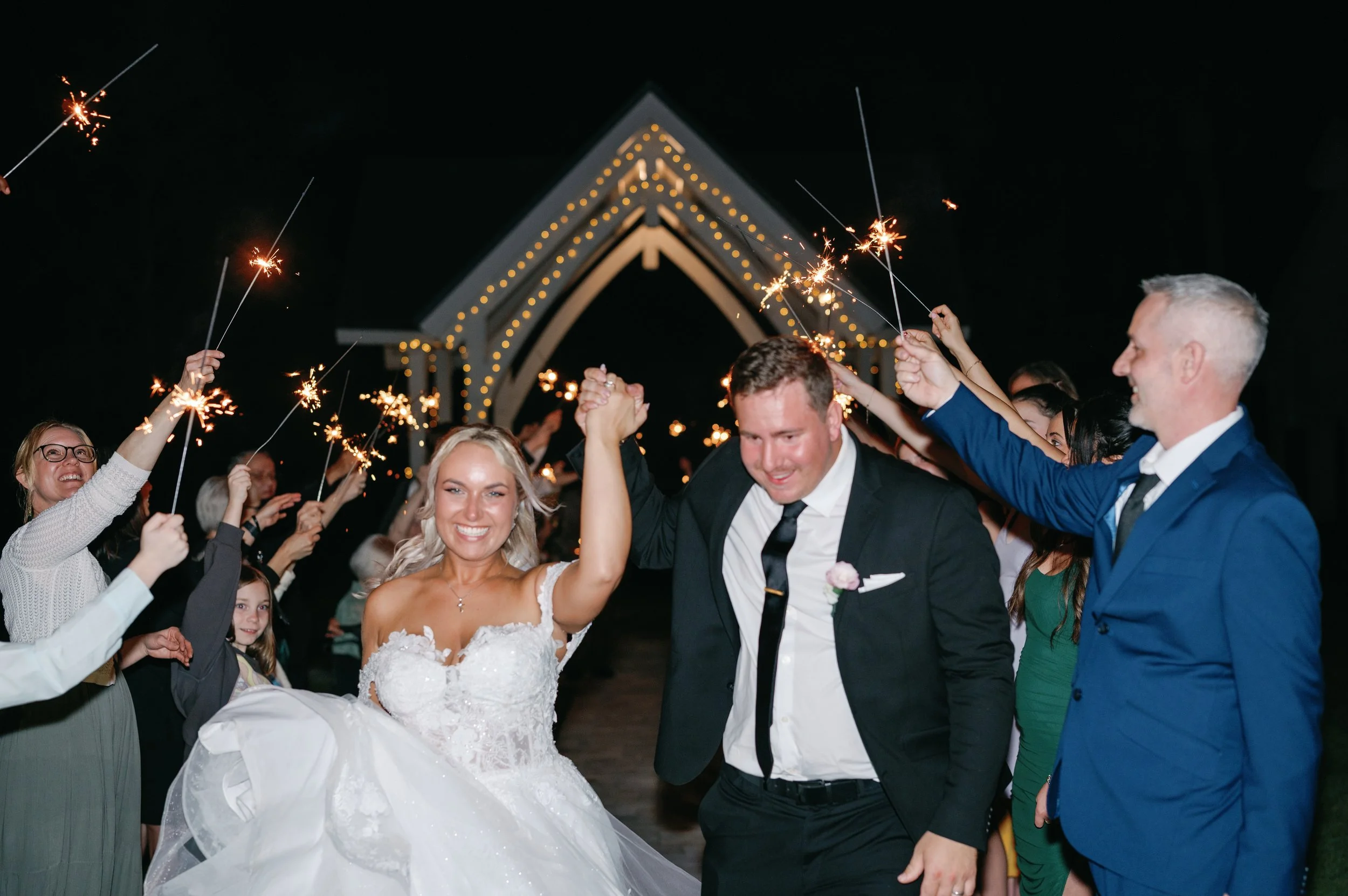 Bride and groom at their wedding reception, holding hands and smiling as guests celebrate with sparklers outdoor at night.
