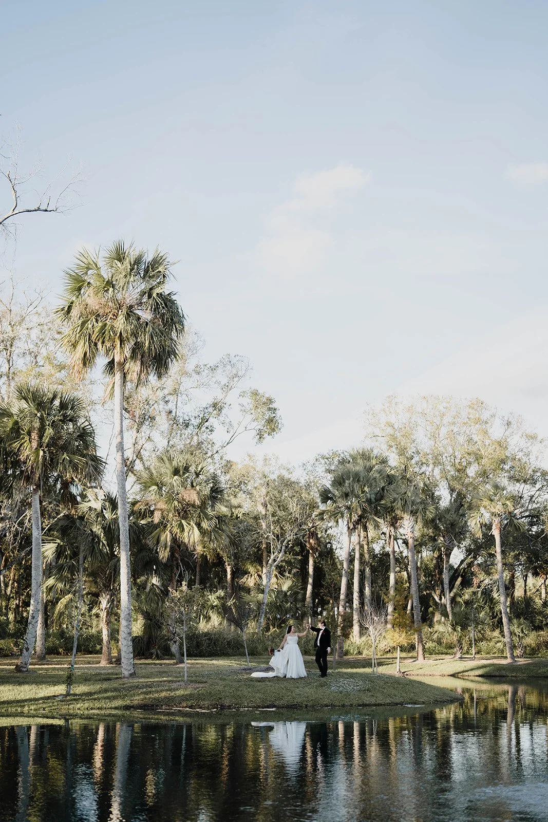 A bride and groom standing on a grassy patch near a body of water, surrounded by tall palm trees and other greenery, with a light blue sky overhead.