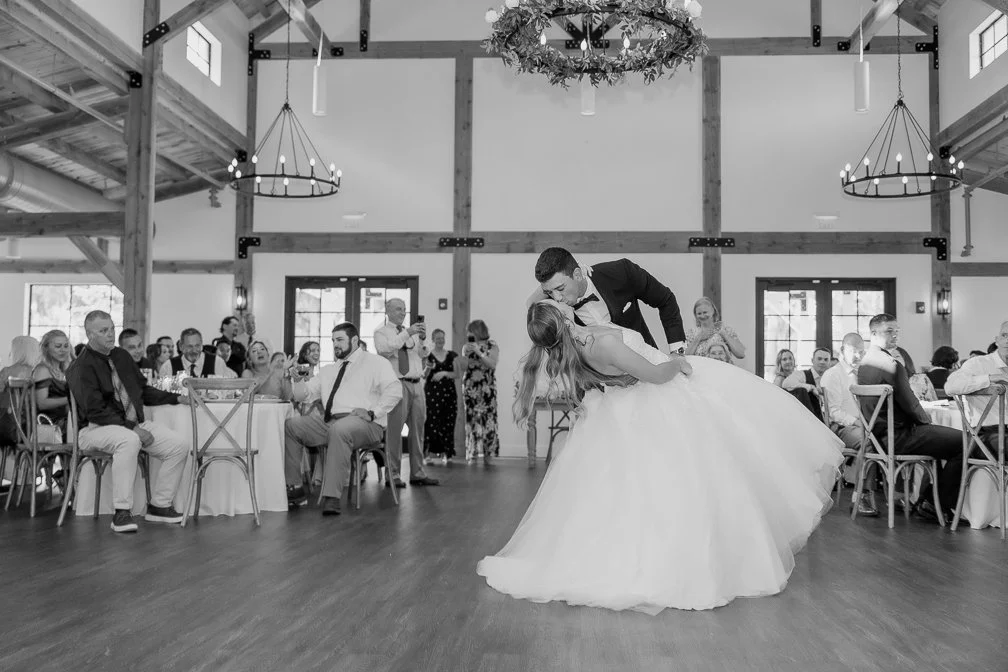 Black and white photo of a bride and groom dancing at their wedding reception in a rustic barn setting, with guests seated around tables watching them.