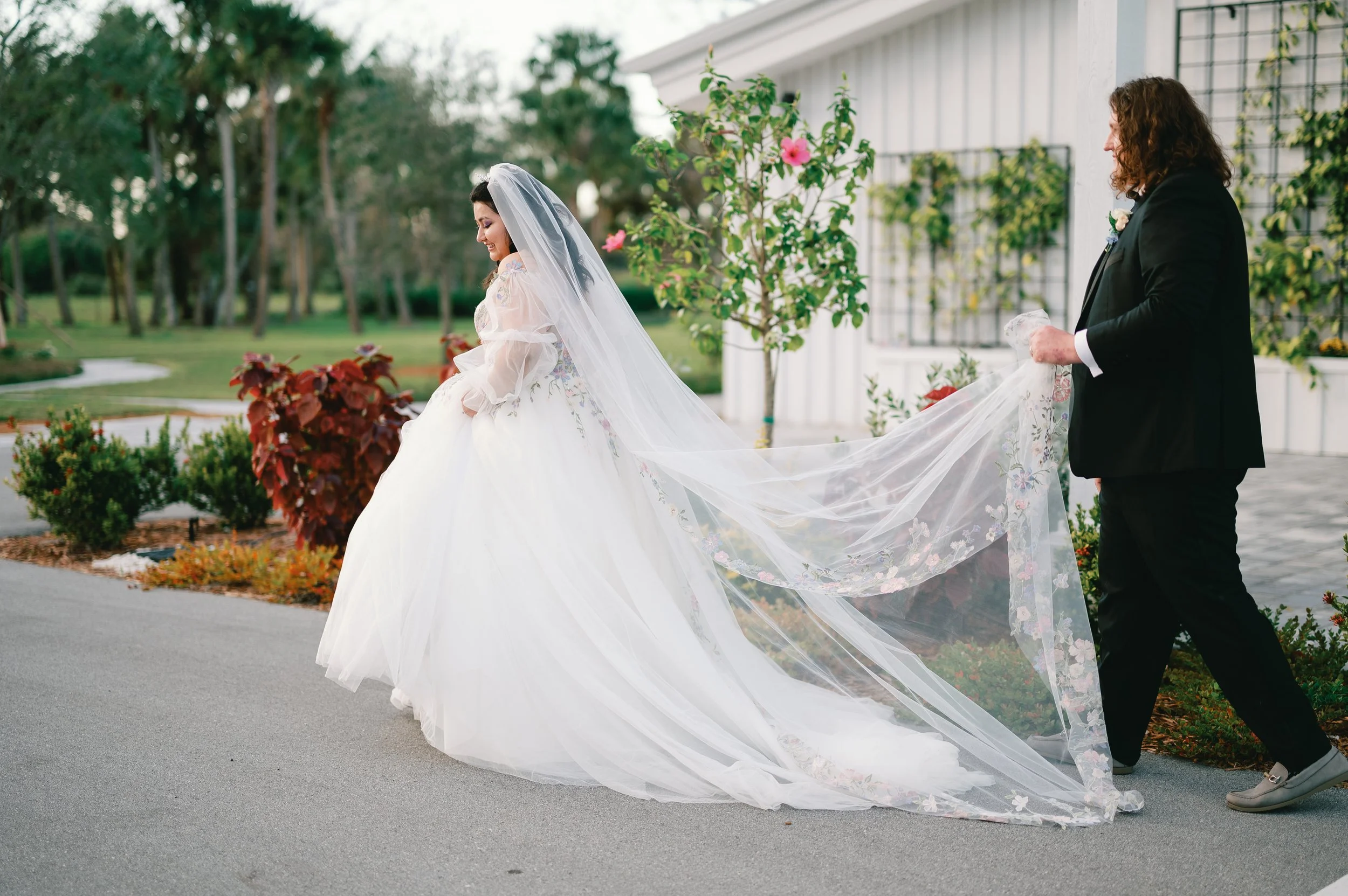 A bride in a white wedding dress with a long train and veil stands outside, smiling, as a groom in a black suit holds the train.