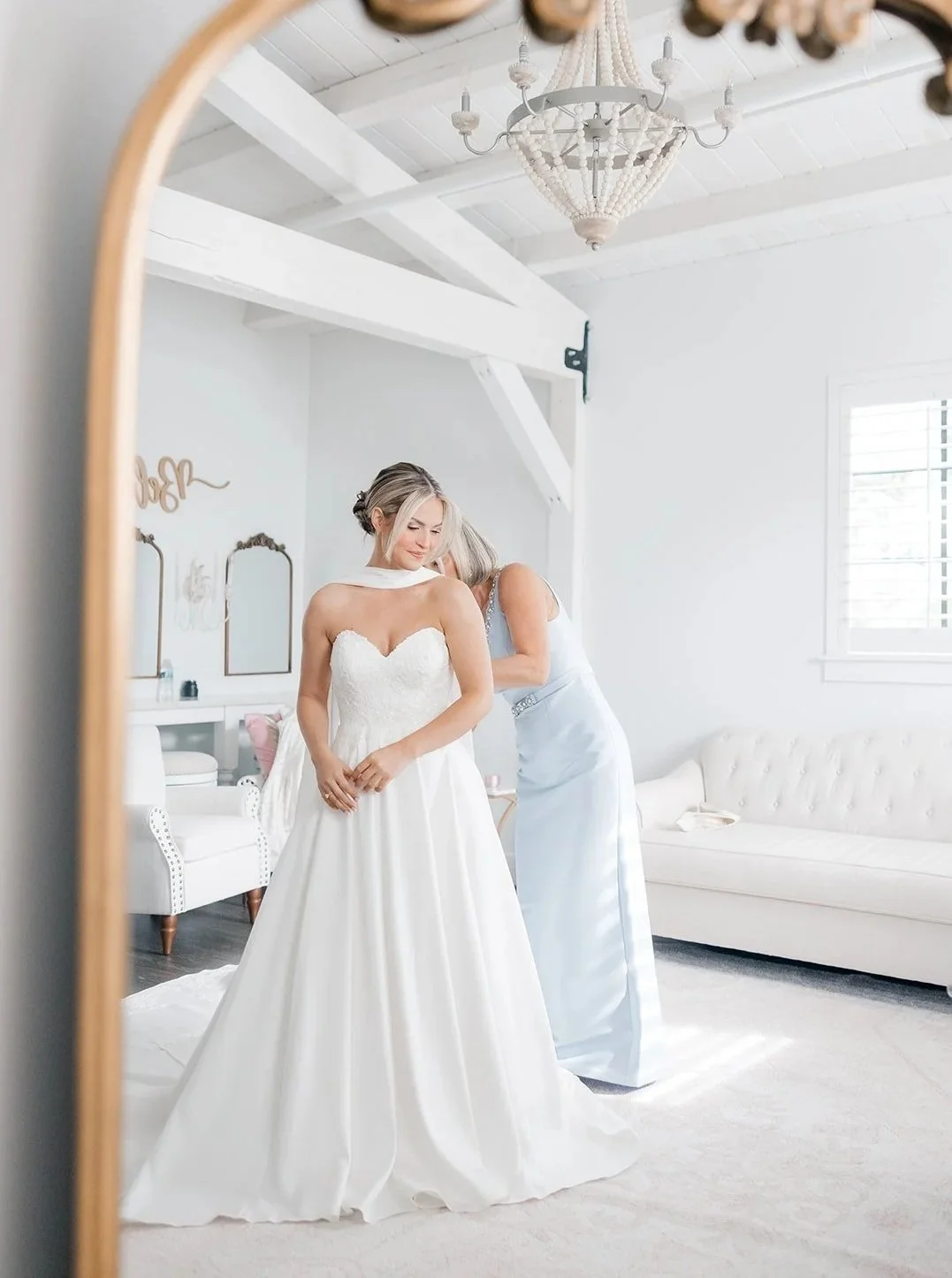 A bride in a white wedding dress is getting ready, standing in front of a mirror while a woman helps her with her dress in a bright, white room with natural light, a white sofa, and chandelier.