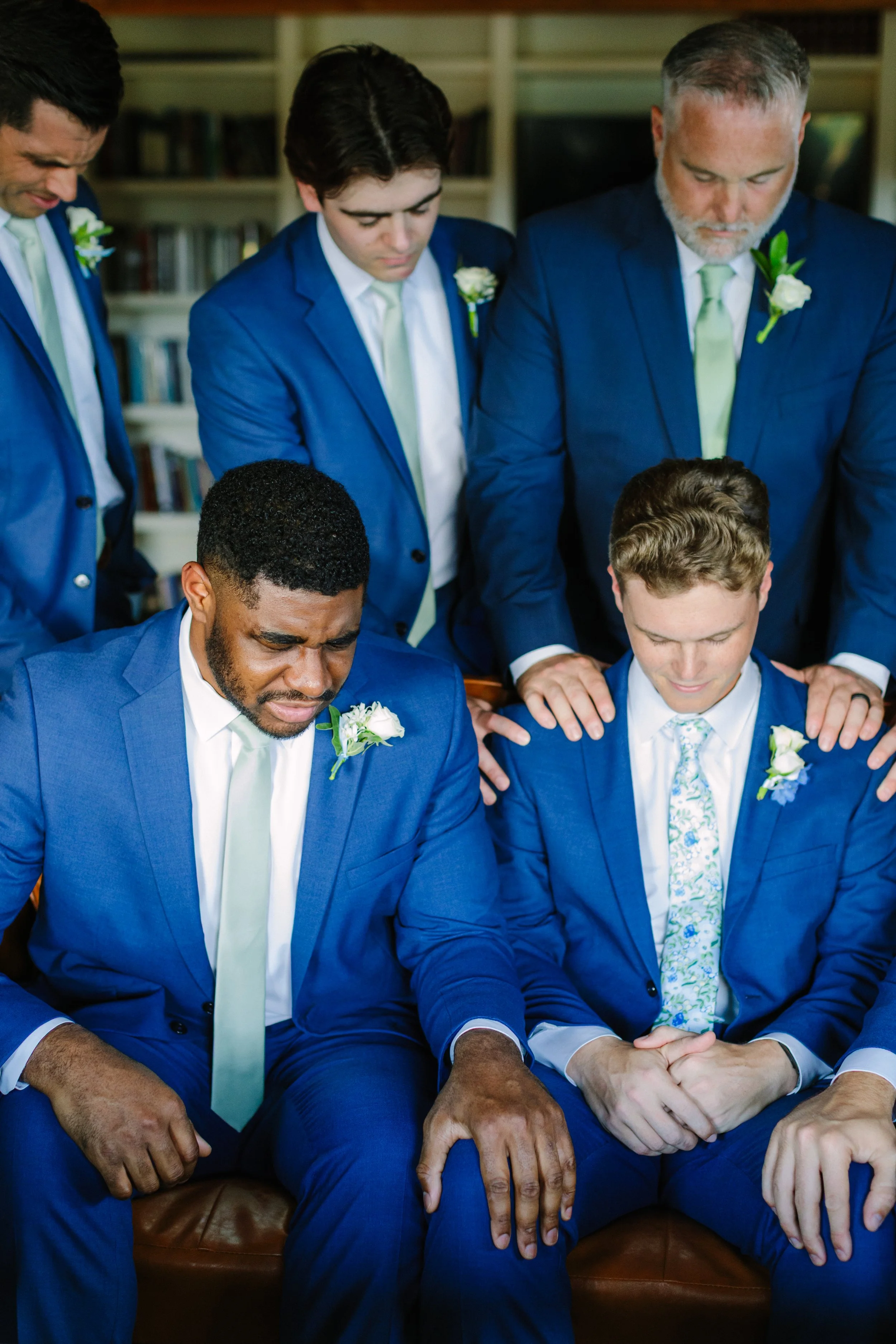 Group of six men in blue suits participating in a prayer or moment of reflection, with two seated men bowing their heads and four standing behind them with hands on their shoulders, in an indoor setting with bookshelves in the background.