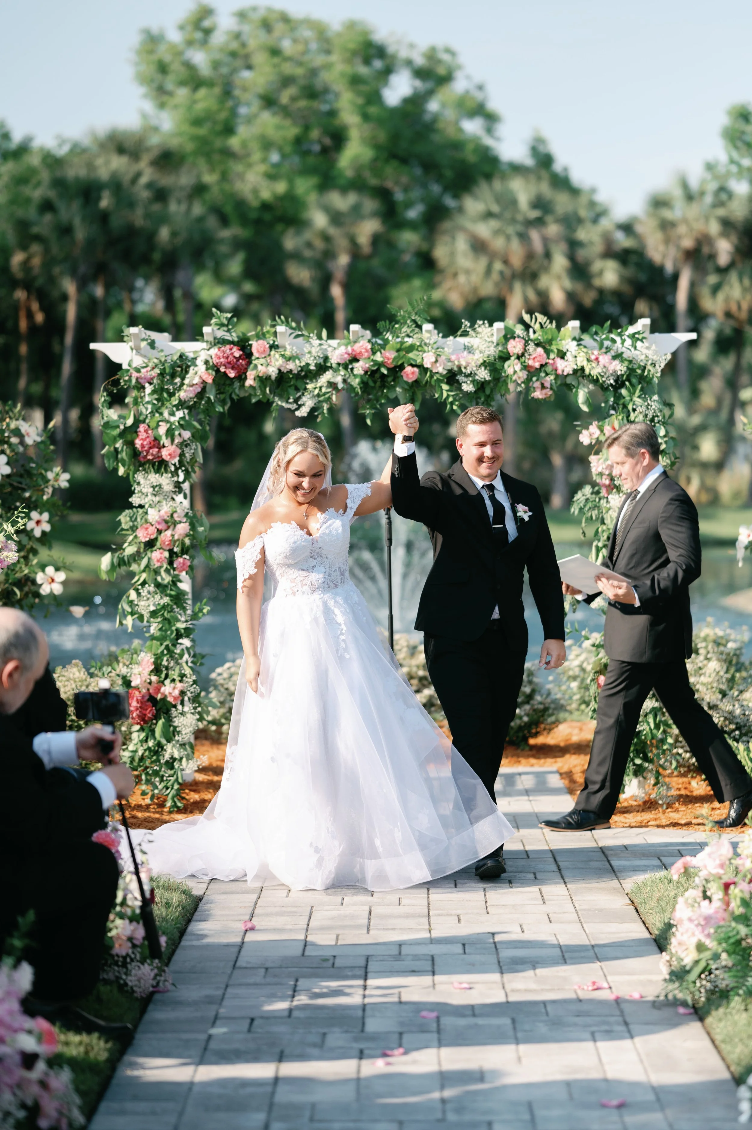 A newlywed couple celebrating their wedding outside under a floral arch, with the bride in a white gown and the groom in a black suit, holding their hands up in victory as an officiant looks on and a photographer captures the moment.