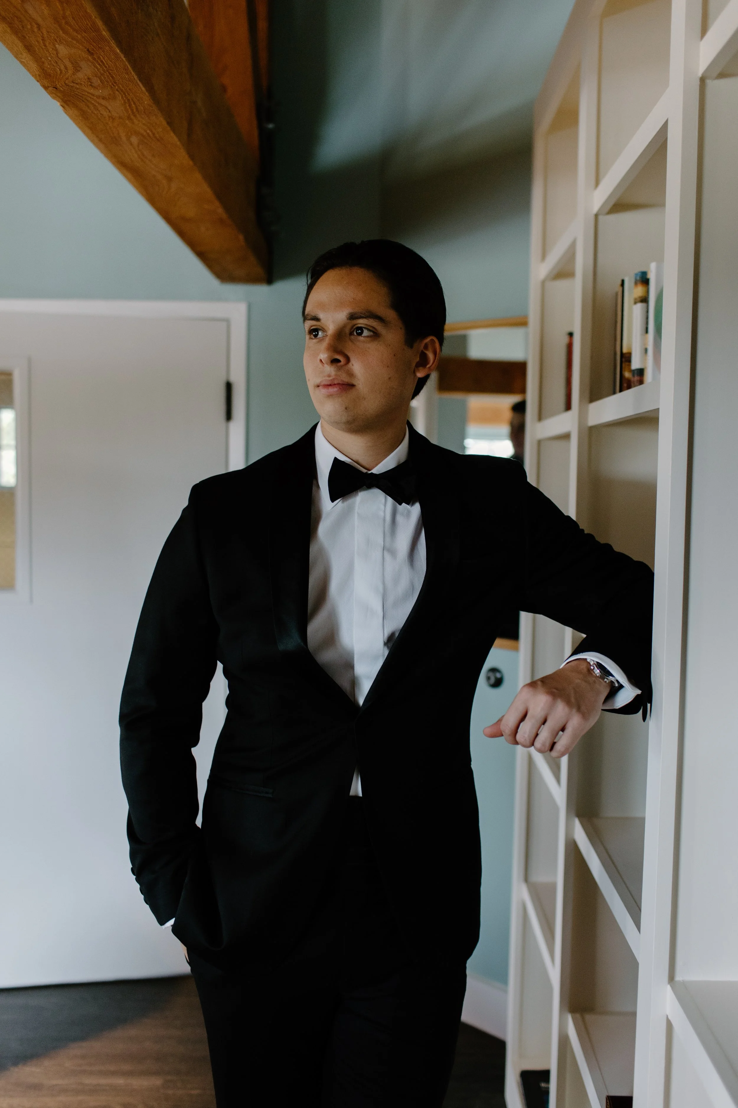 A young man in a black tuxedo with a bow tie standing beside a white bookshelf in a room with wooden beams and a door.