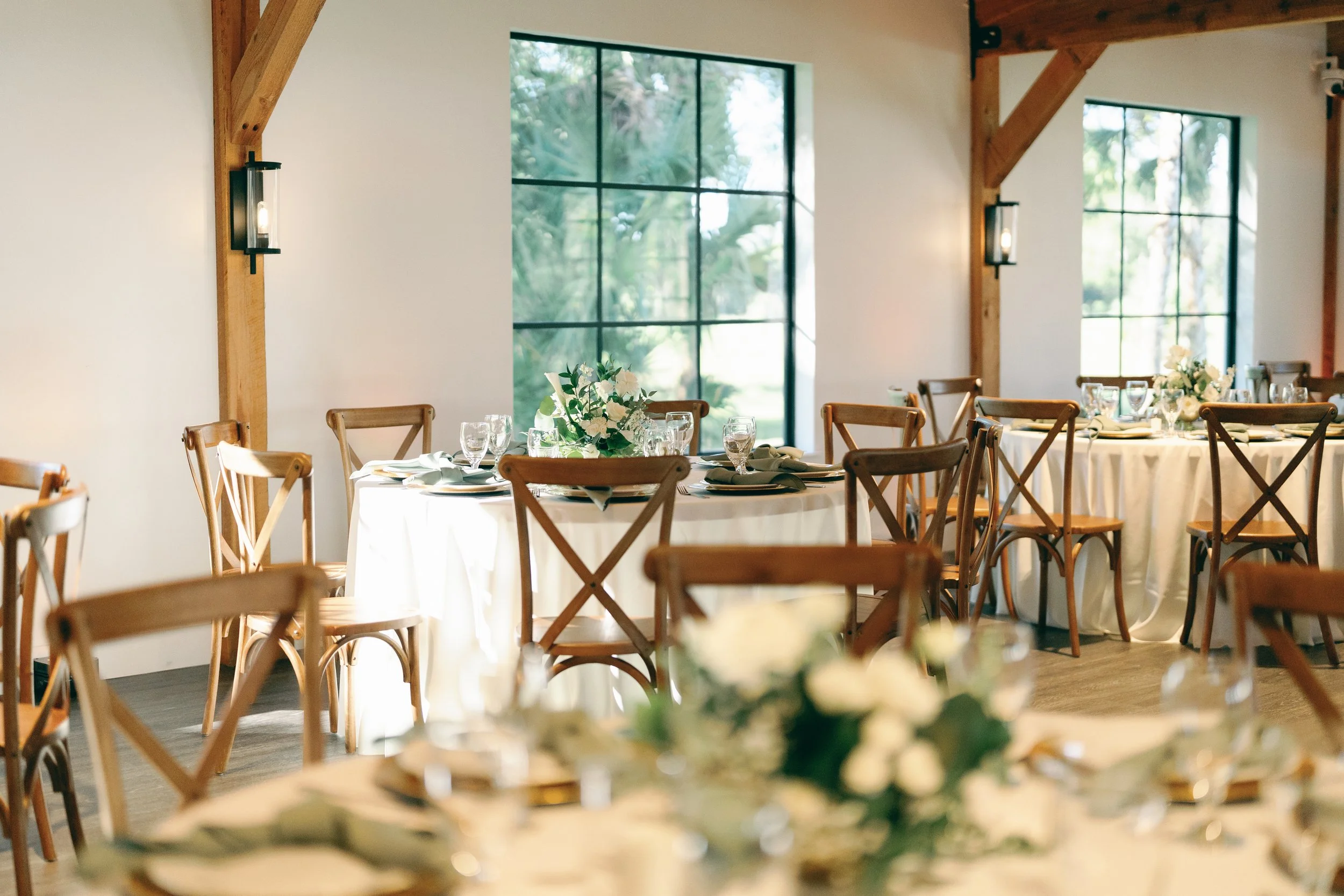 Elegantly set banquet hall with round tables covered in white tablecloths, decorated with floral centerpieces, and surrounded by wooden chairs. Large windows with black frames allow natural light to illuminate the room. Wooden beams and modern wall s