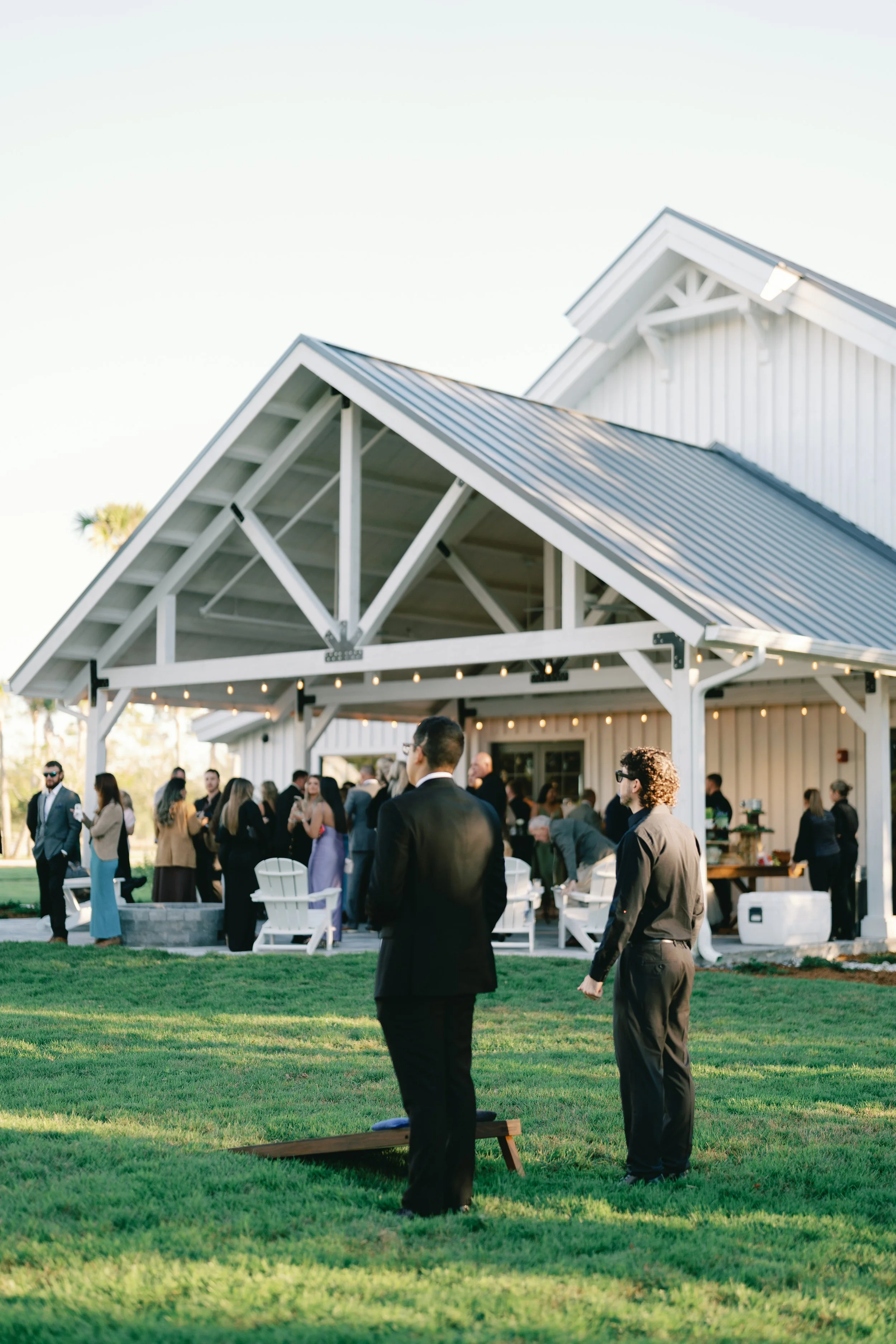 A gathering of people at an outdoor event under a white barn with string lights, with two men in suits standing in the foreground on a grass lawn.