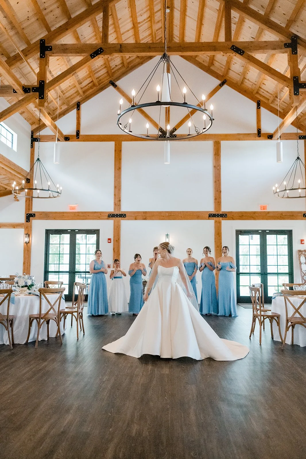 Bride in a white gown dancing while her bridesmaids in blue dresses watch in a rustic, wooden wedding venue with high vaulted ceilings and chandeliers.