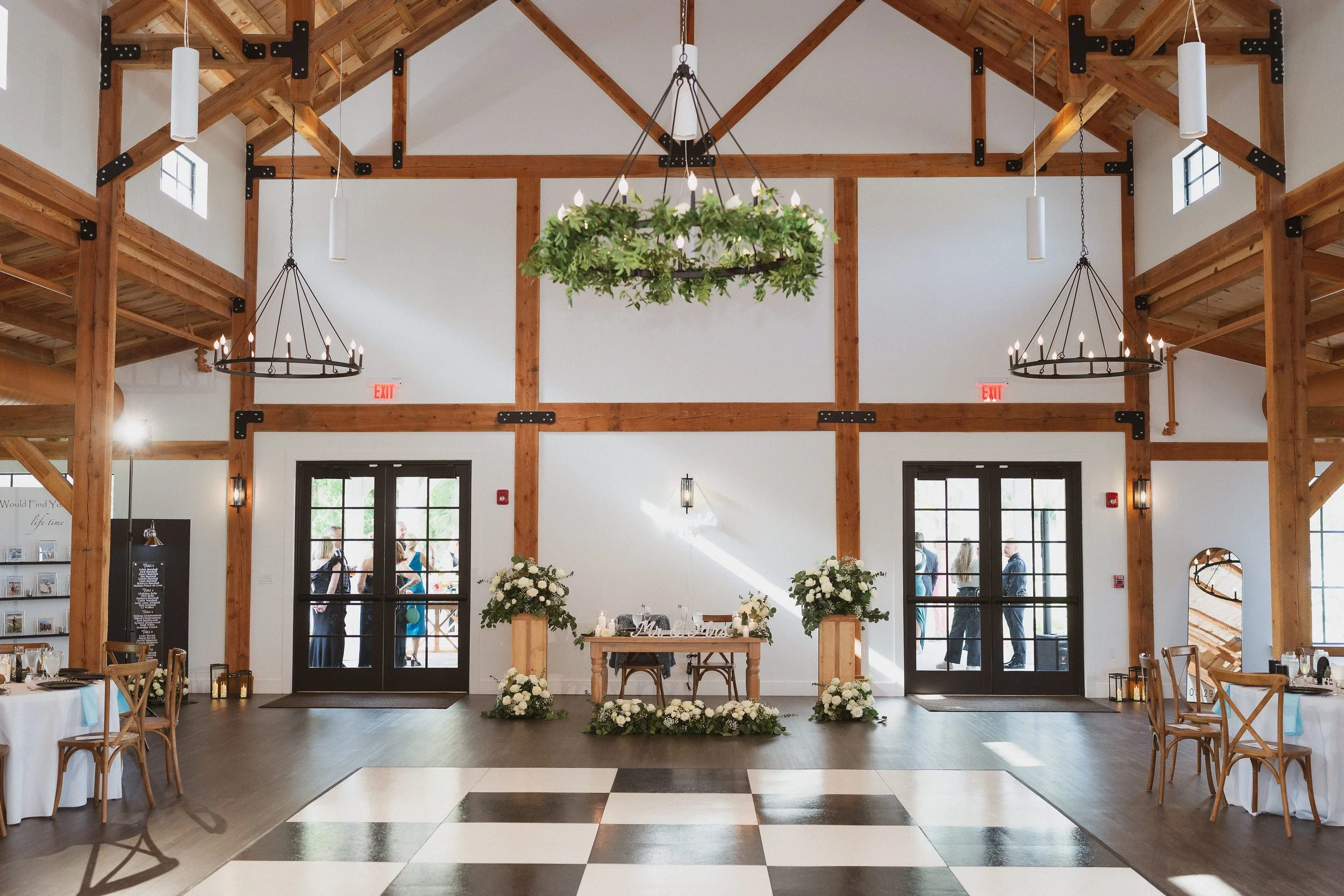 Interior of a rustic wedding reception hall with high wooden beams, chandeliers, floral arrangements, and a checkered dance floor in the center.