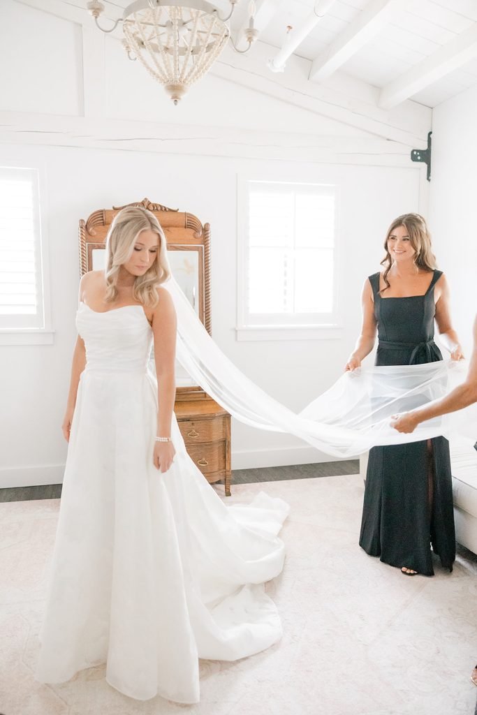 A bride in a white wedding dress standing in the bridal suite at Celebration Grove with two windows, while a woman in a black dress helps adjust her veil.