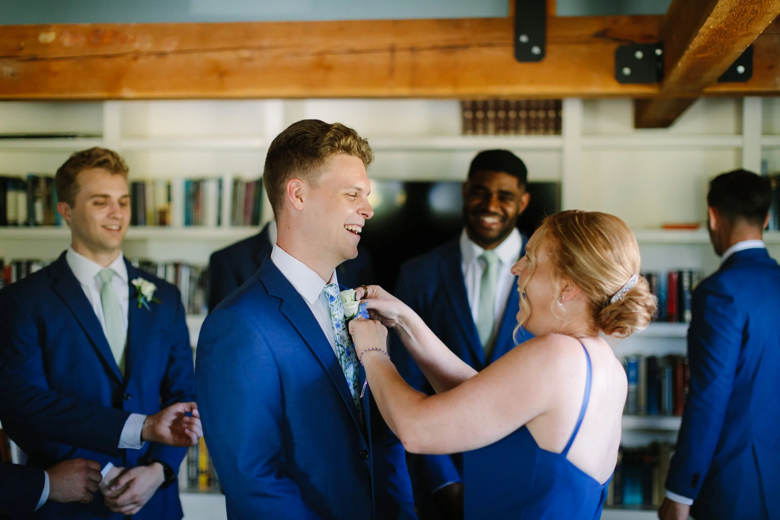 A woman pins a boutonniere on a groom's suit as part of a wedding ceremony. The groom is smiling and surrounded by groomsmen dressed in matching blue suits.