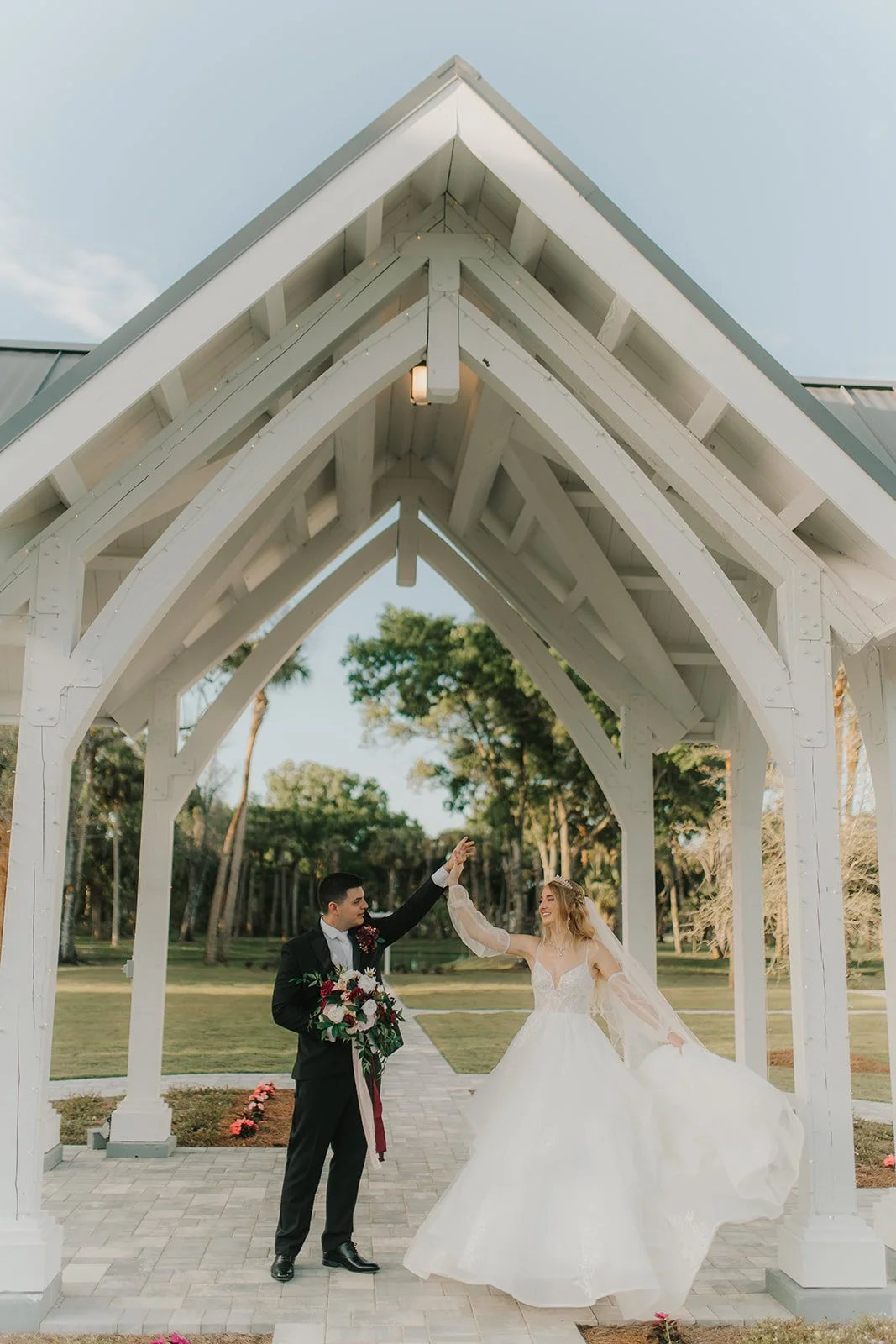 Groom and bride dancing under a white wooden wedding arch outside, with greenery and trees in the background.
