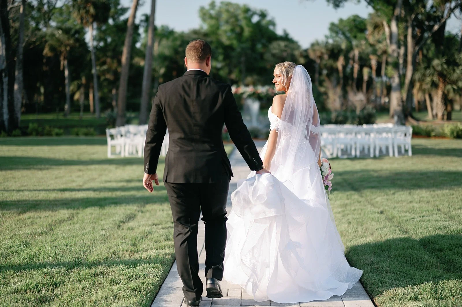 Bride and groom walking hand in hand toward the Lakeside Arbor at Celebration Grove, featuring fountain lake views, outdoor ceremony seating, and Florida’s ancient oaks in Vero Beach.