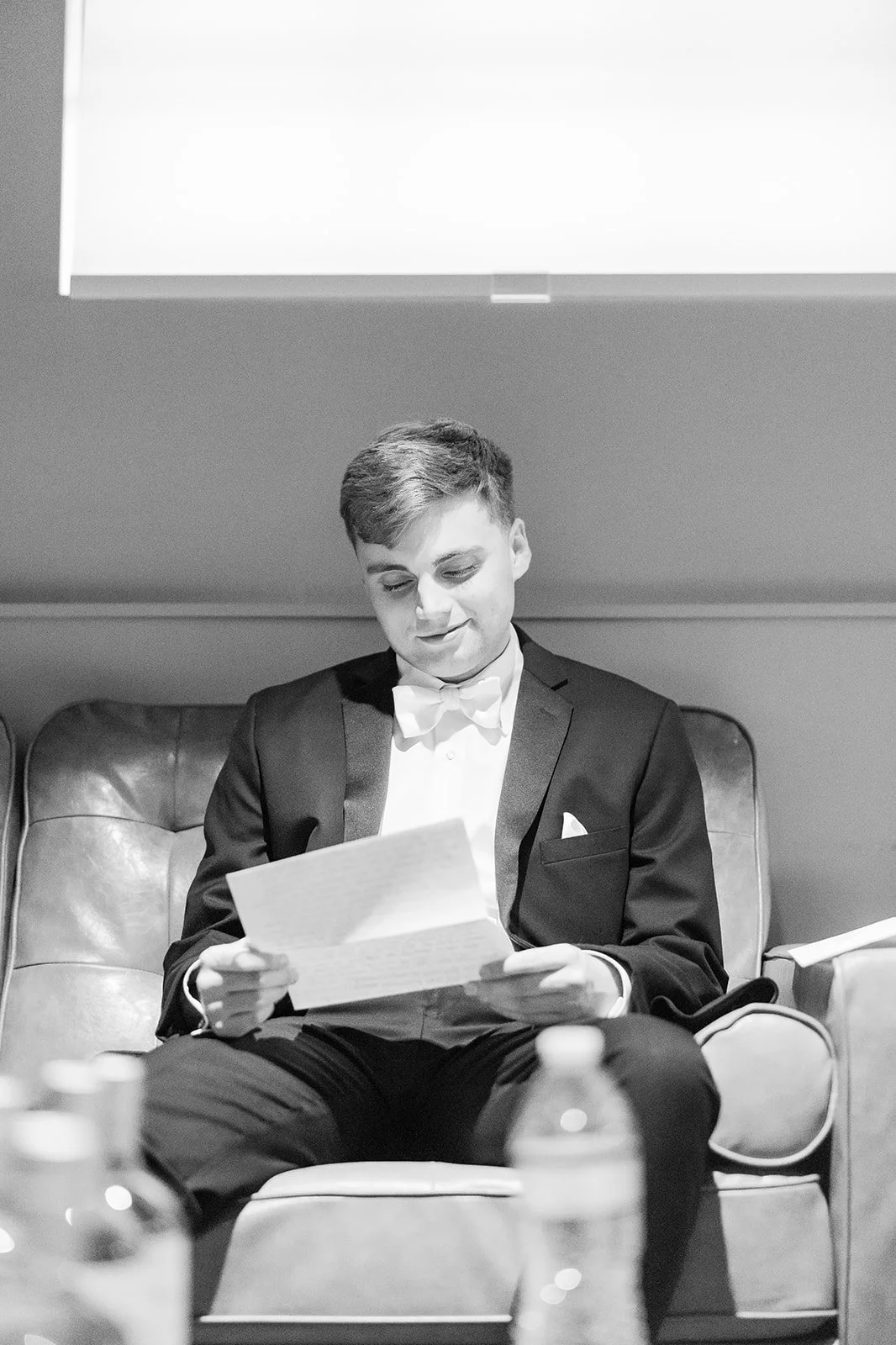 A young man in a tuxedo sitting on a leather couch, smiling and reading a piece of paper, with bottles on the table in front of him.