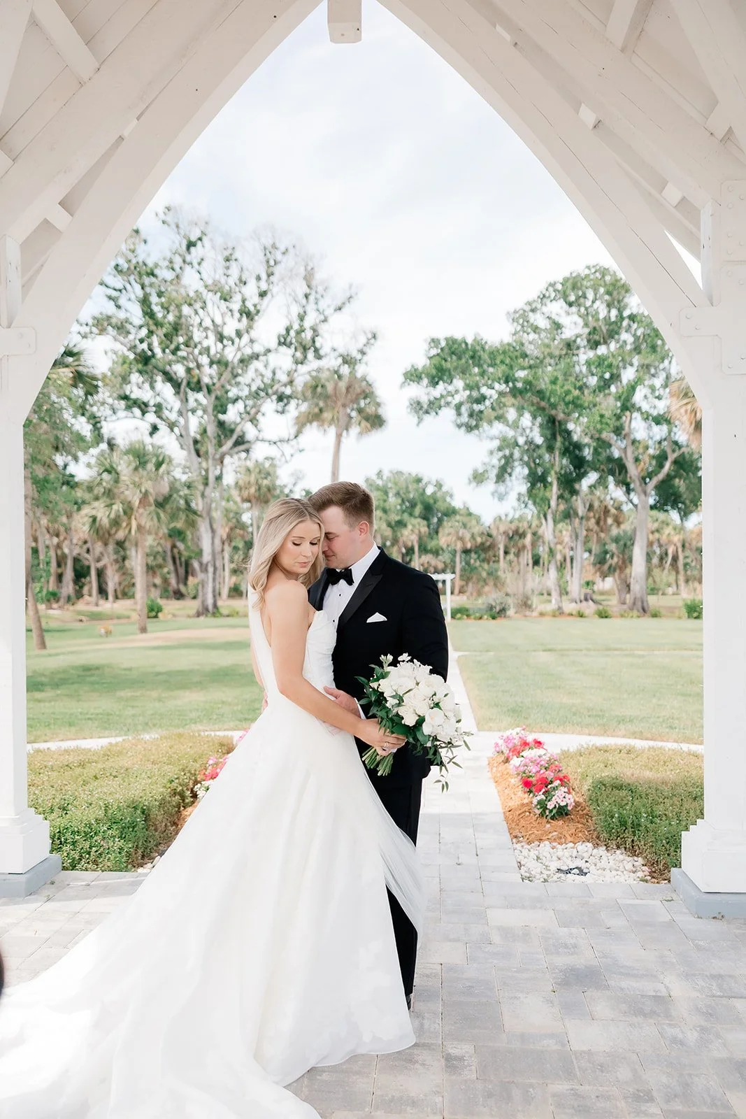 Bride and groom standing close together under a white pavilion, holding a bouquet of white flowers, with trees and a pathway in the background.