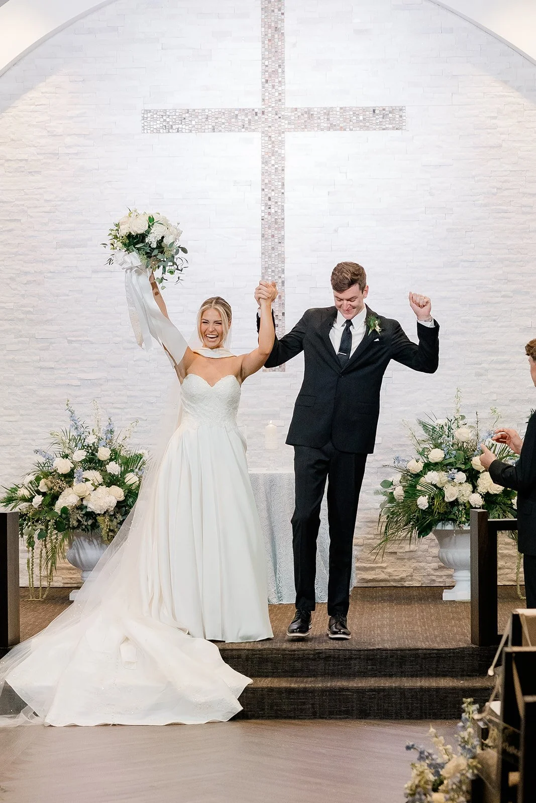 A bride and groom celebrate after getting married in a church, holding hands and smiling with arms raised. The bride is wearing a white wedding dress and the groom is in a black suit. There are floral arrangements and a cross on the wall behind them.