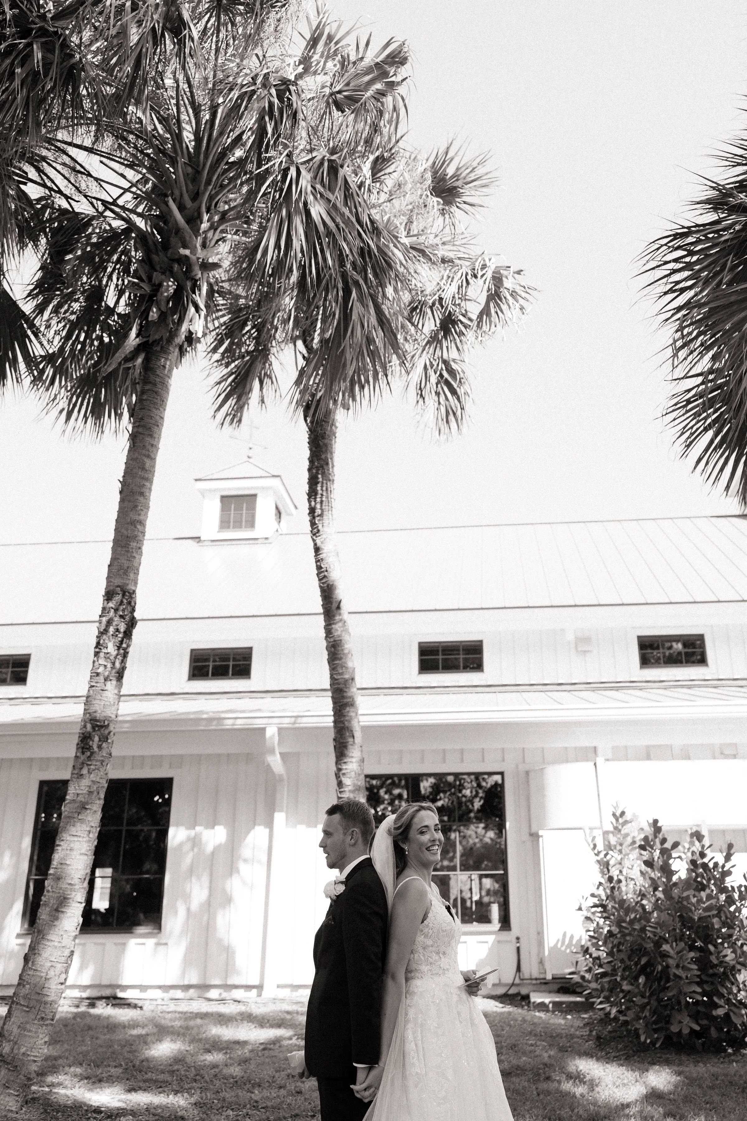 A black and white photo of a bride and groom holding hands back-to-back outdoors, in front of a house with large windows and a metal roof, near two tall palm trees.