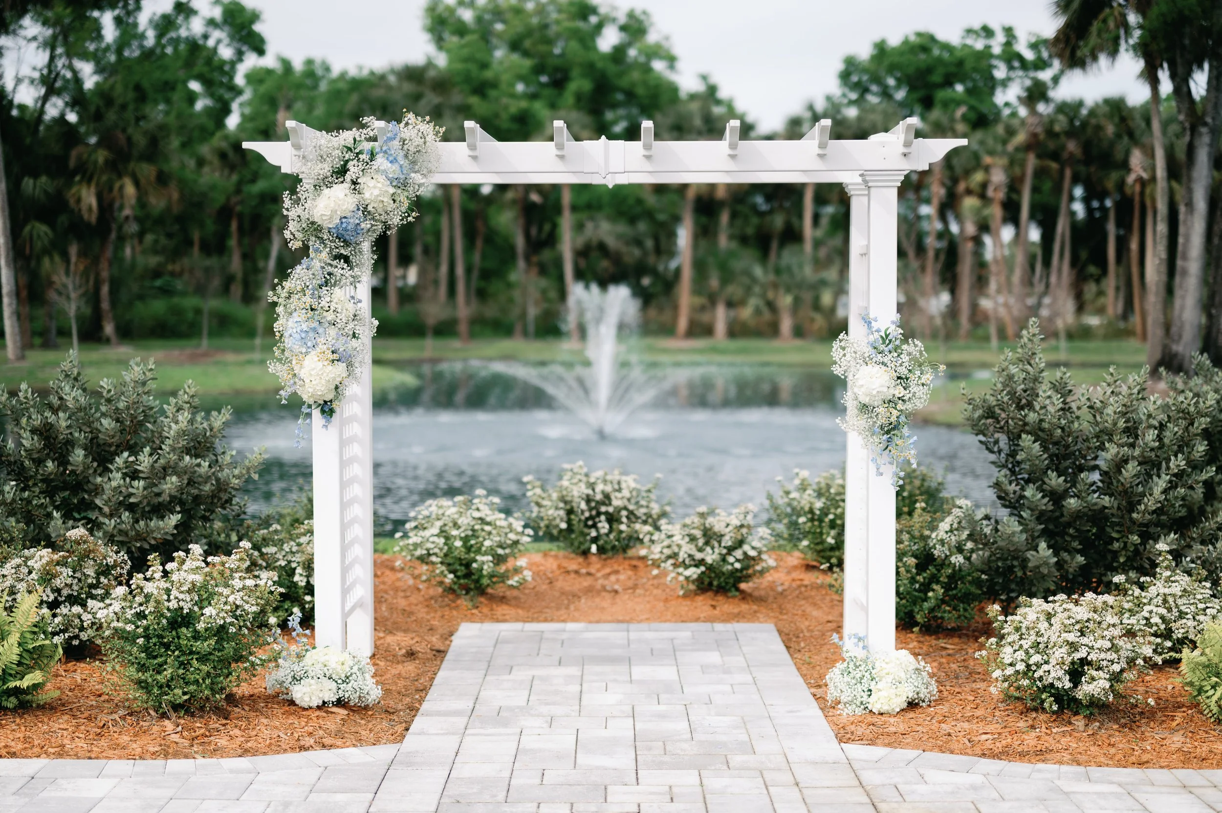 A white wedding arch decorated with white and blue flowers, set over a paved area near a pond with a fountain, surrounded by green bushes and trees.