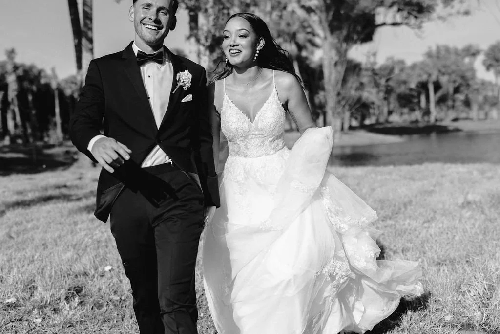 Black and white photo of a smiling bride and groom walking outdoors on grass, with trees and water in the background. The groom is in a tuxedo, and the bride is in a wedding dress, holding up the skirt as they walk.