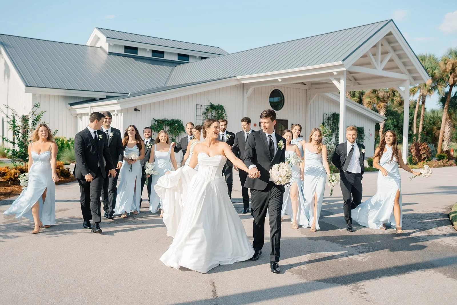 A bride and groom leading a wedding procession of bridesmaids and groomsmen outside a white church on a sunny day.