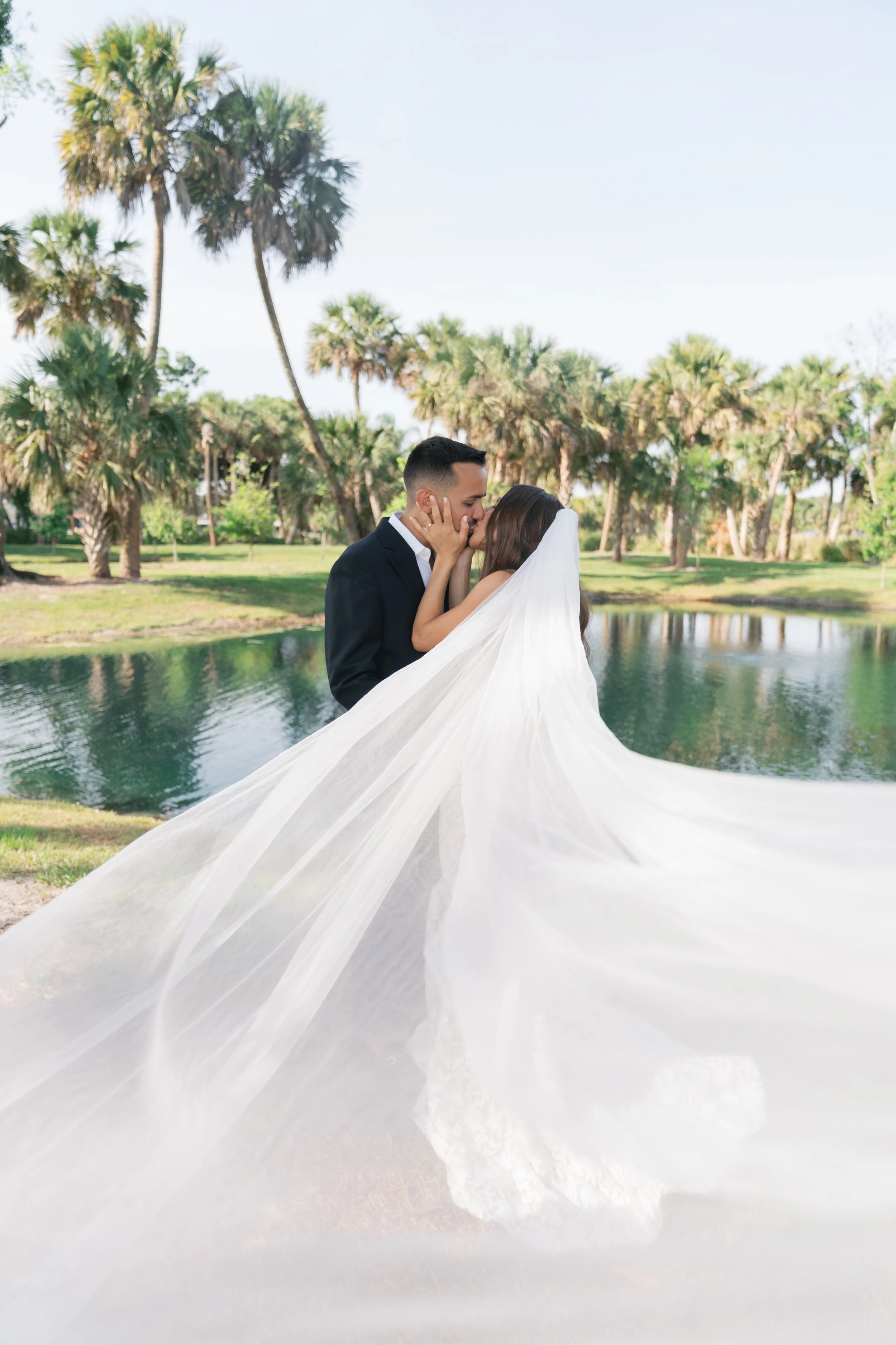 Bride and groom sharing a kiss outdoors near a lake with palm trees in the background, the bride's white veil flowing in the wind.