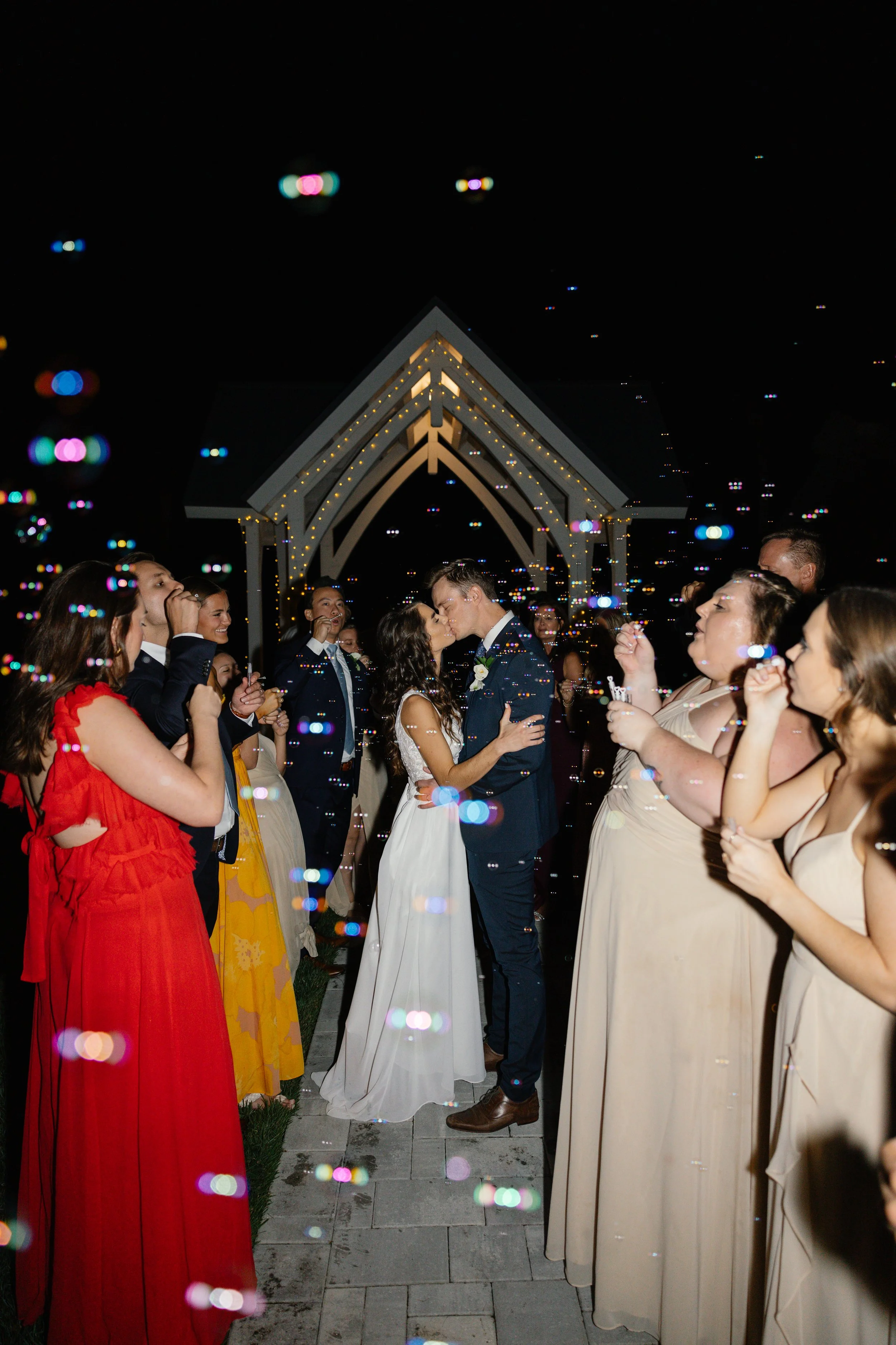 A wedding celebration at night with a bride and groom kissing under a decorated arch, surrounded by guests blowing bubbles.