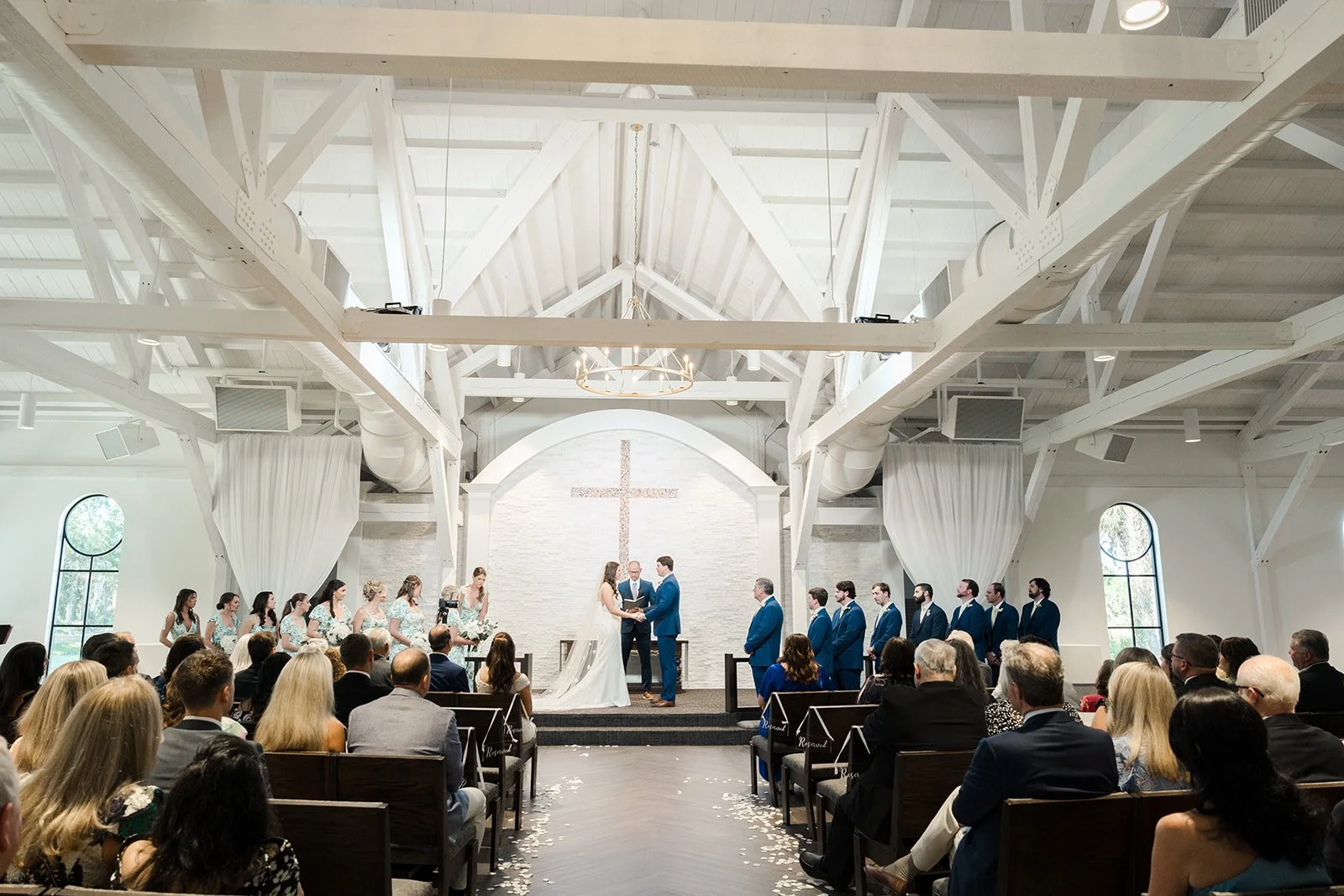 A wedding ceremony inside a church with white wooden beams, a cross on the wall, and guests seated on wooden pews. The bride and groom are at the altar exchanging vows, with bridesmaids and groomsmen standing nearby.