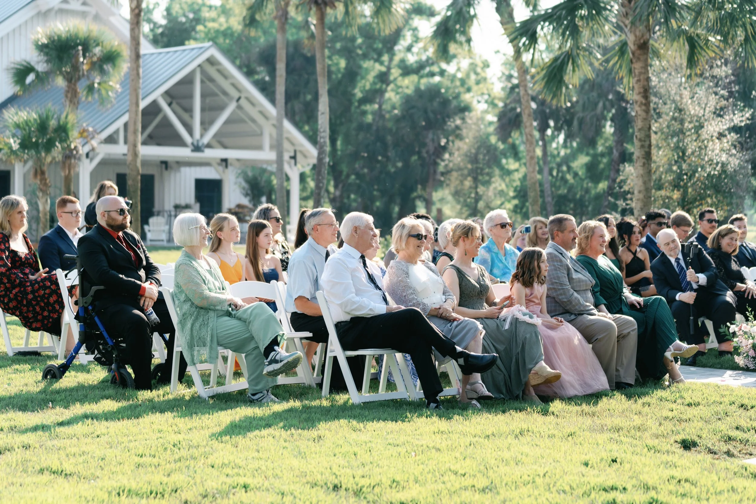 Attendees seated outdoors at a wedding ceremony, with a white house and palm trees in the background.