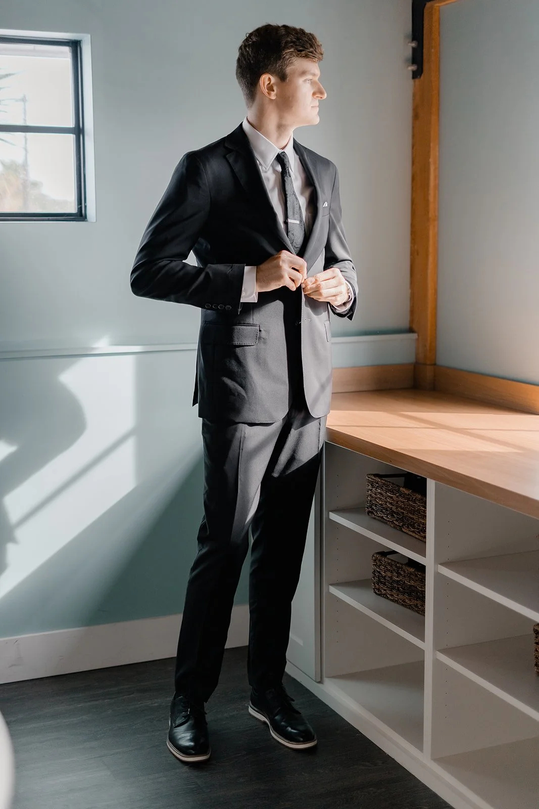 A man in a black suit standing next to a wooden counter, adjusting his jacket, looking out the window.