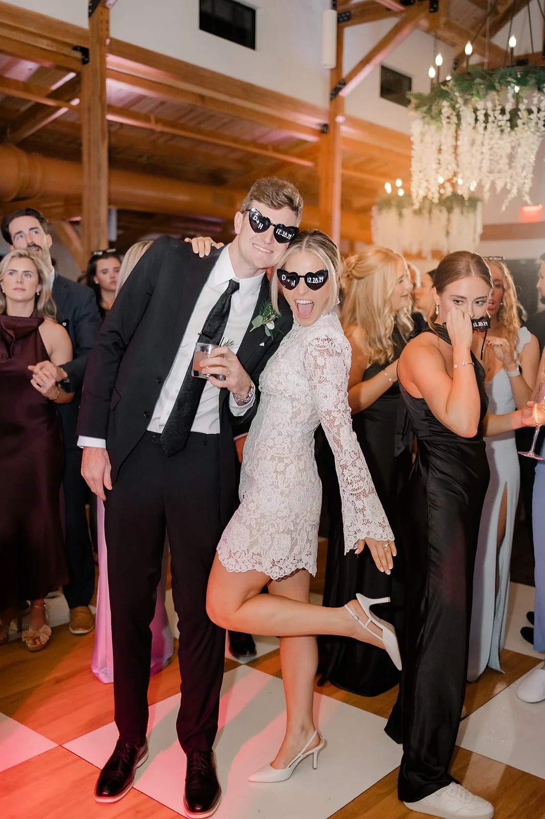 People celebrating at a wedding reception, with two individuals wearing sunglasses with the date 12.25.21, in a decorated venue.