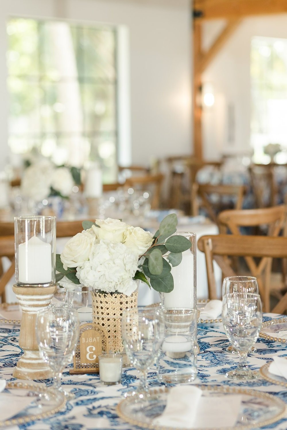 Elegant table setting with white floral centerpiece, candles, and glassware, decorated for a wedding or event in a bright, rustic venue with wooden chairs.