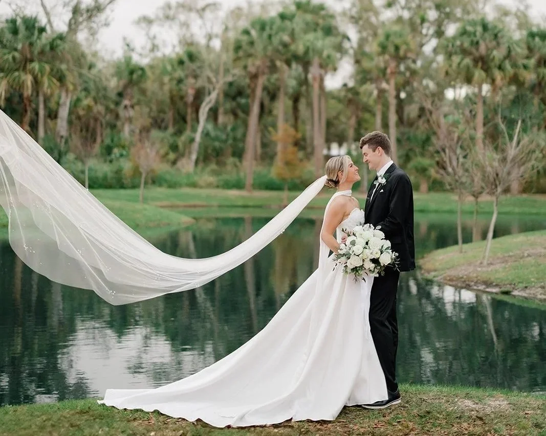 Golden hour wedding portraits at Celebration Grove fountain lake in Vero Beach with flowing bridal veil, ancient oaks, and sunset reflections.