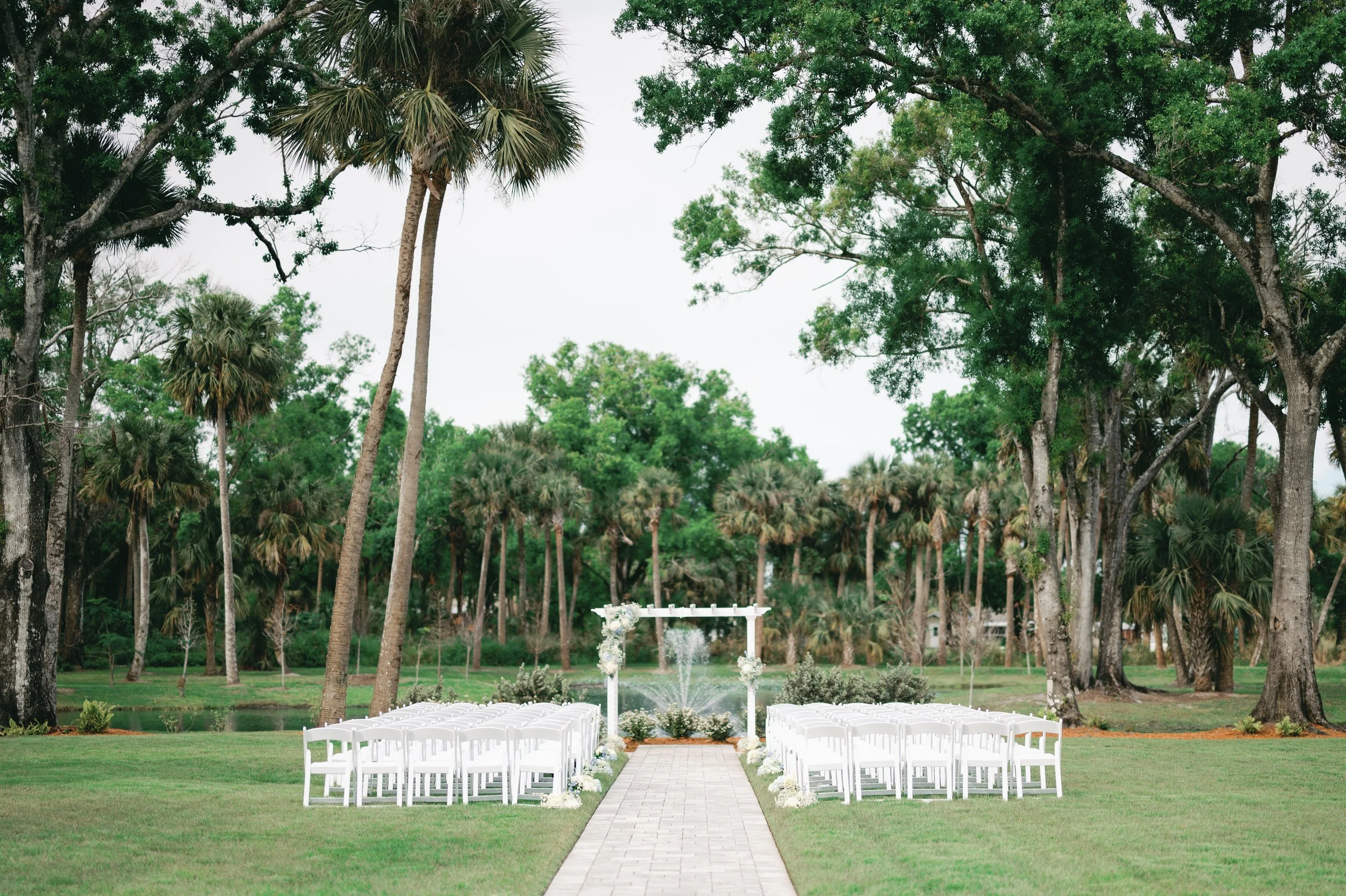 Outdoor wedding ceremony setup with white chairs arranged in two sections on either side of a paved aisle leading to a decorated arch with flowers and a fountain, surrounded by tall trees and palm trees on a lush green lawn.