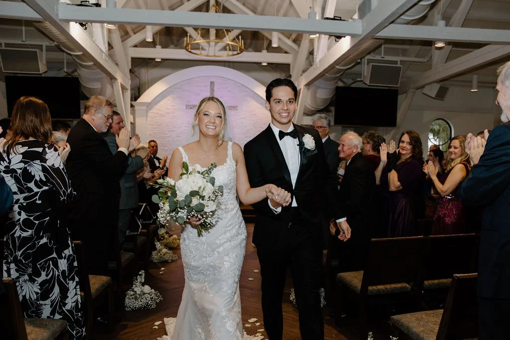 Bride and groom walking down the aisle at their wedding ceremony, holding hands, with guests clapping and smiling around them.