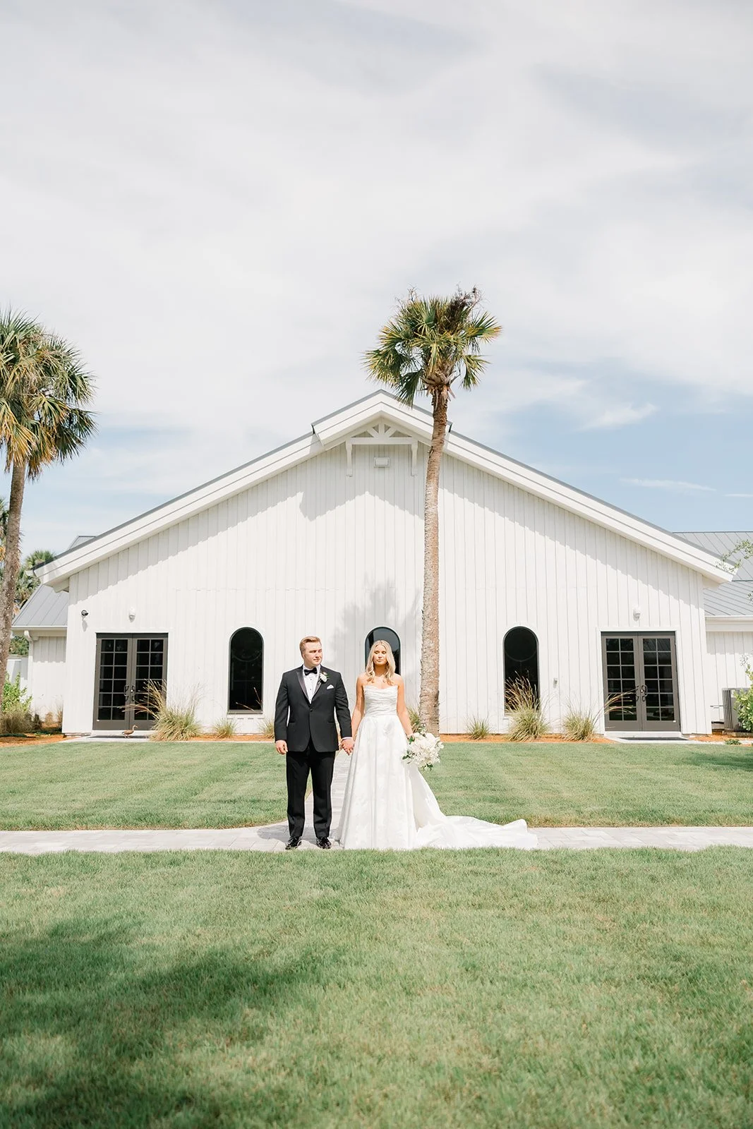A bride and groom holding hands standing in front of a white building with palm trees, green grass, and a partly cloudy sky.