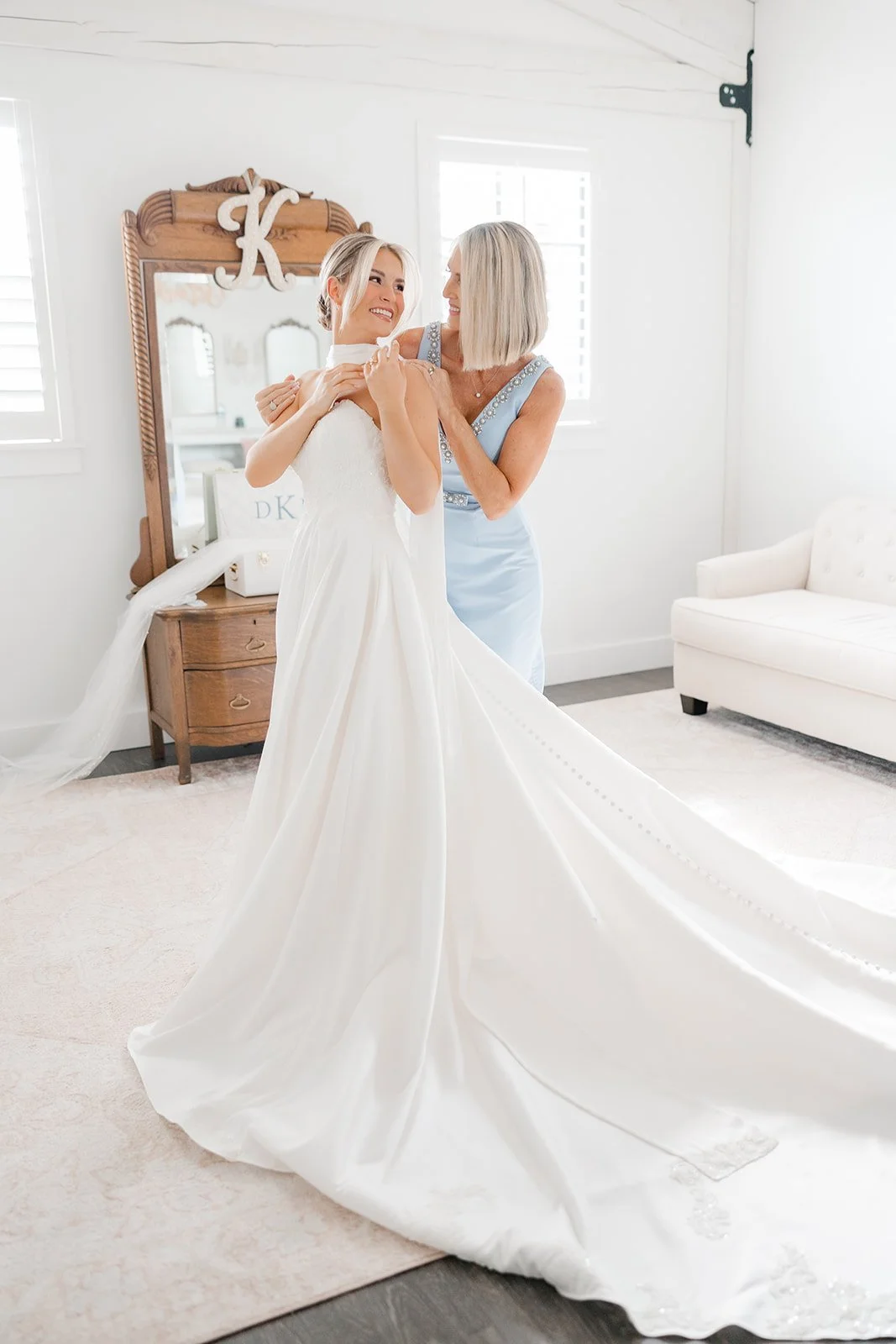 A bride in a white wedding dress is being helped by a woman in a light blue dress as they smile at each other in a bright, white room with a mirrored dresser and a beige sofa.
