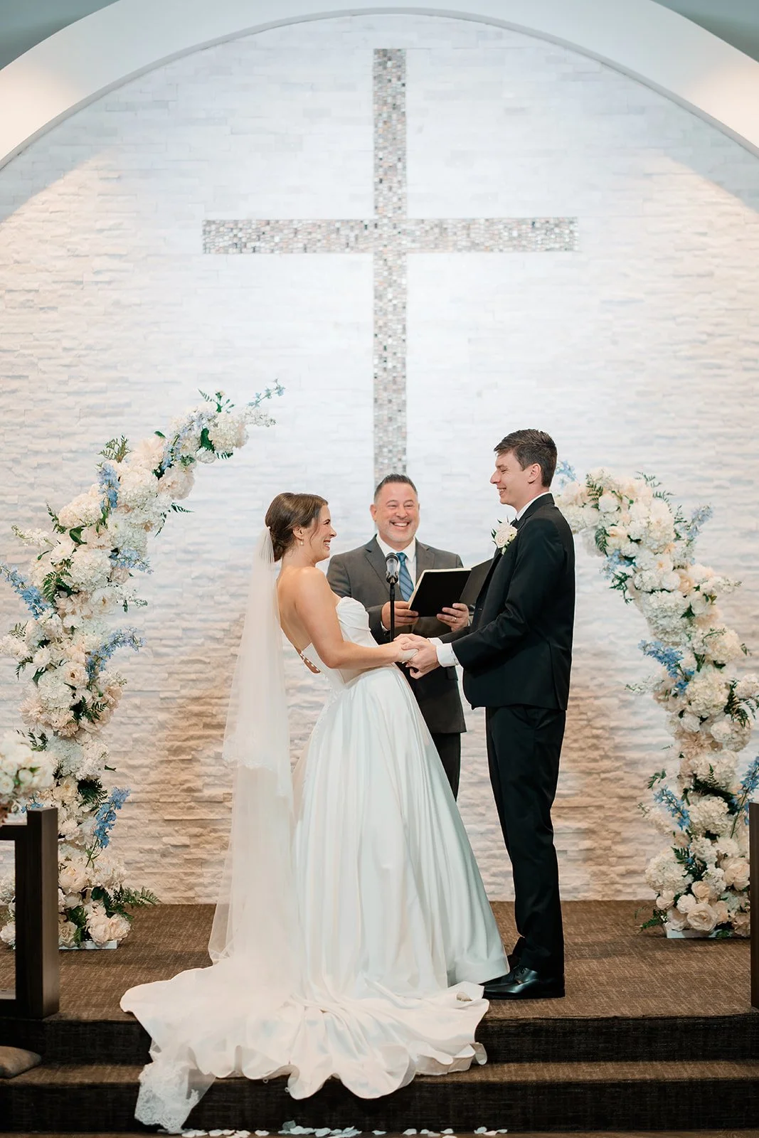 A wedding ceremony with a bride and groom standing face to face, holding hands, and exchanging vows in front of a white brick wall with a large cross. The officiant, standing behind them with a microphone, is smiling. The scene is decorated with whit