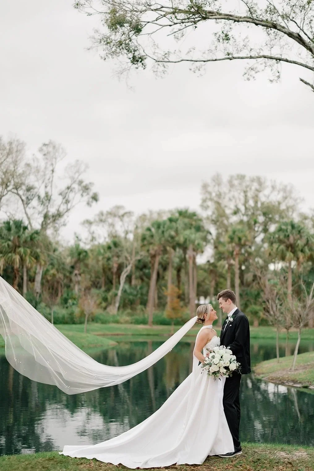 A bride and groom stand close together by a lake during their wedding at Celebration Grove, with the bride holding a bouquet of white flowers, the bride's veil flowing to the side, and a scenic background of trees and a cloudy sky.
