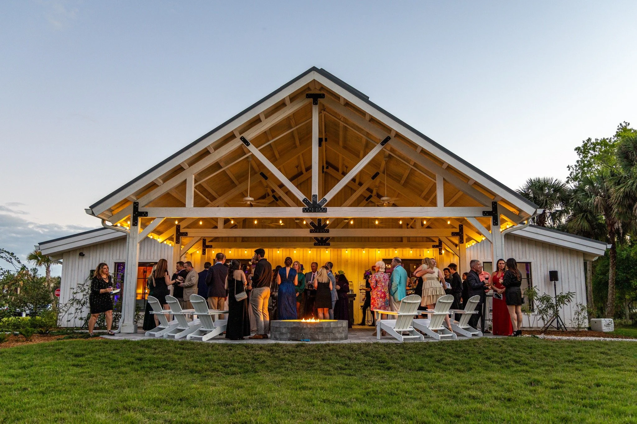 People gathered at an outdoor event under a large open pavilion with a fire pit in front. The pavilion has exposed wooden beams and bright lights, with a grassy area in the foreground and trees in the background.