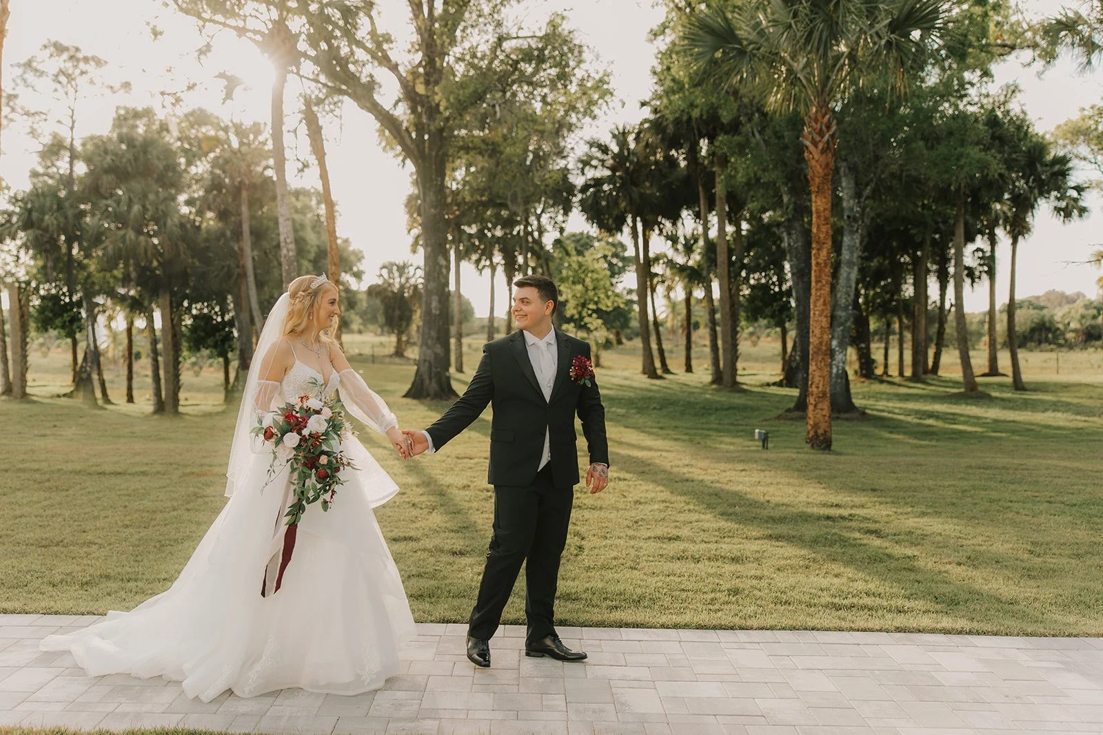 A bride and groom in wedding attire holding hands and smiling at each other outdoors on a sunny day, with trees and greenery in the background.