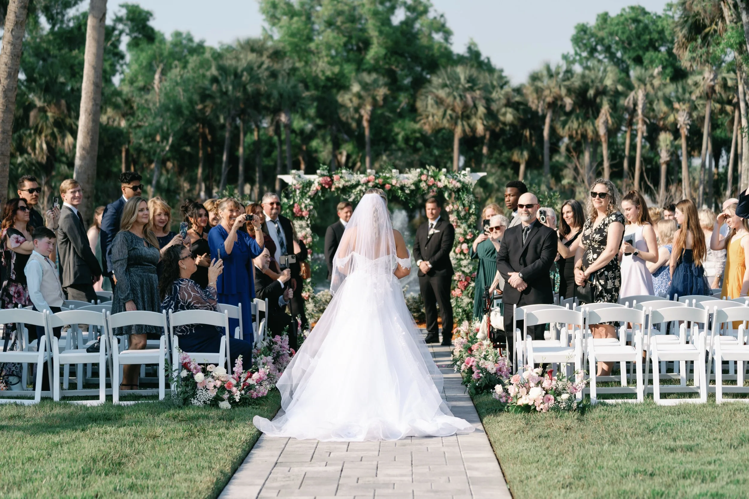 A bride in a white wedding dress and veil stands at the altar during an outdoor wedding ceremony, surrounded by guests, with a floral arch and green trees in the background.